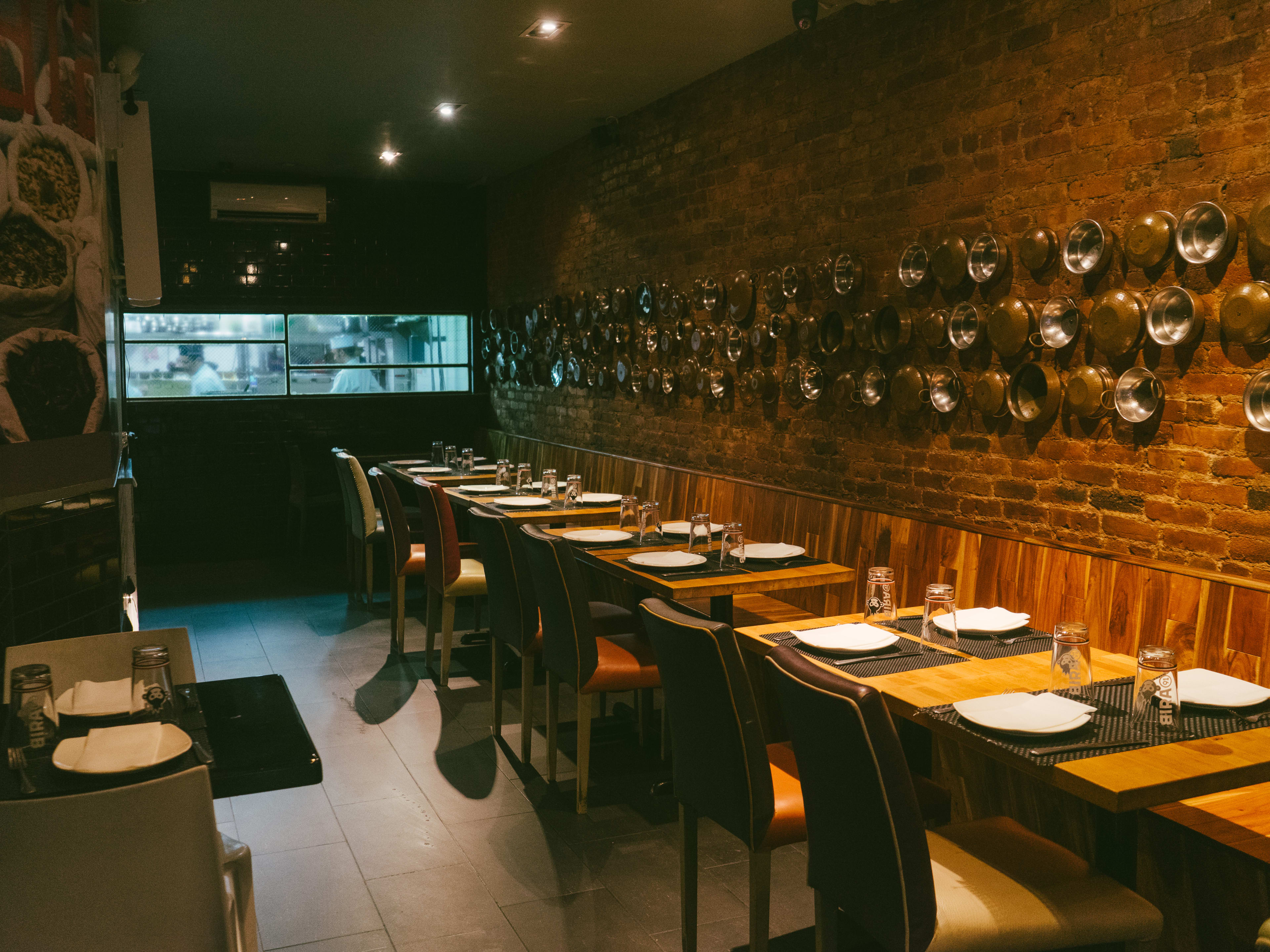 The interior of Malai Marke has copper pots hanging on the brick walls and a long row of booths with tables and chairs. The kitchen is seen through a window in the back.