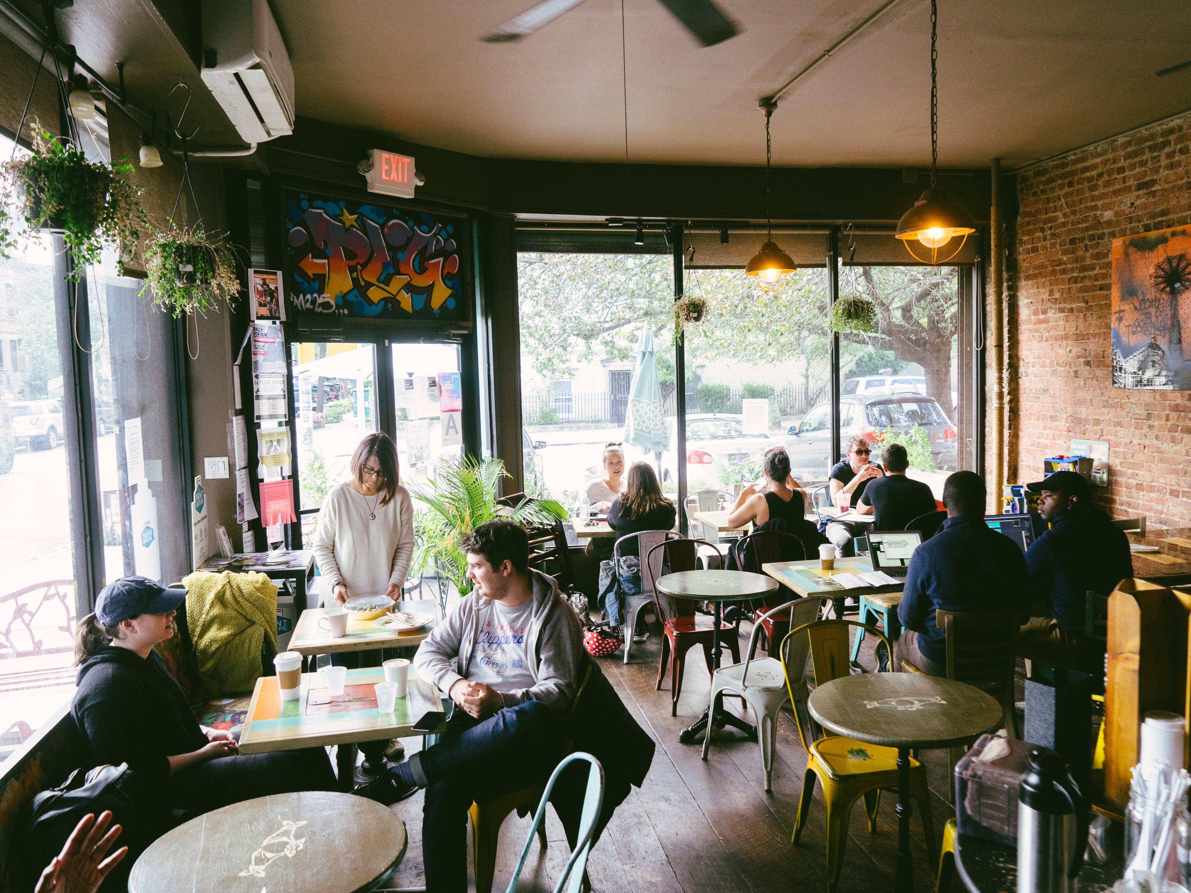 The interior of PLG. There are people at various tables, art on the brick walls and large windows at the front.