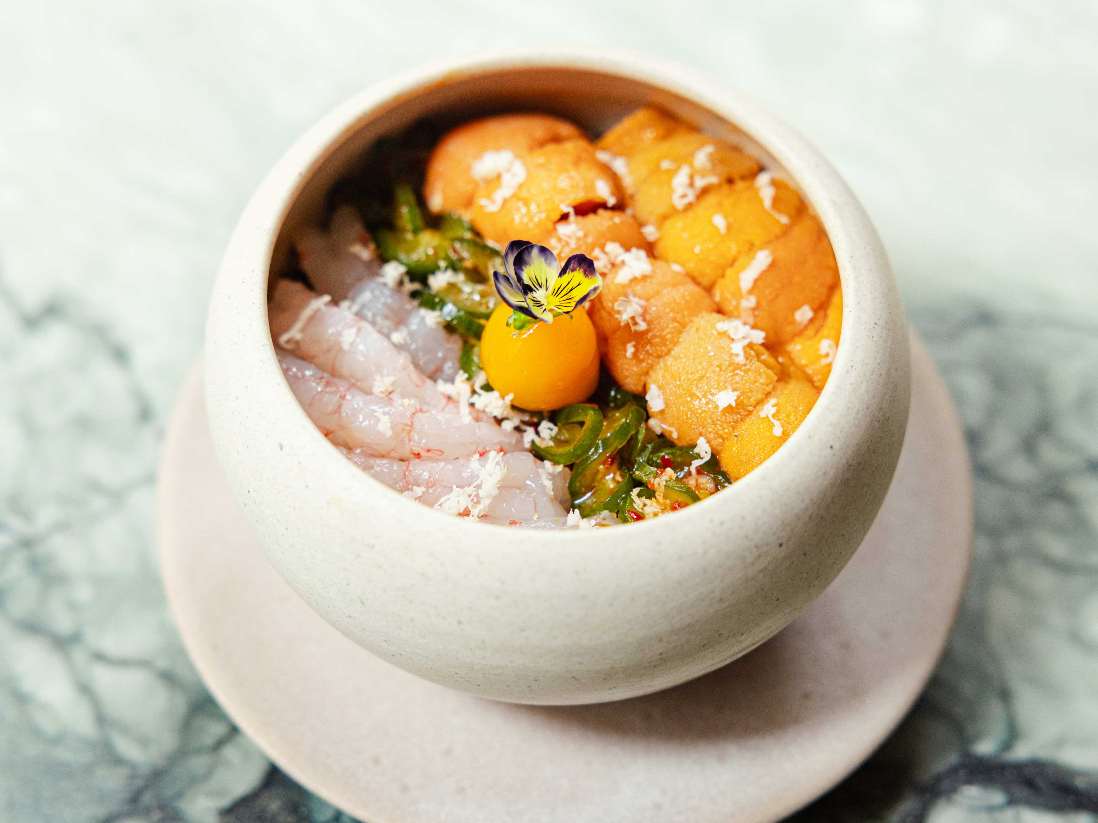 A ceramic bowl with uni, sweet shrimp, spicy cucumber salad, an egg yolk, and a purple flower, over rice.