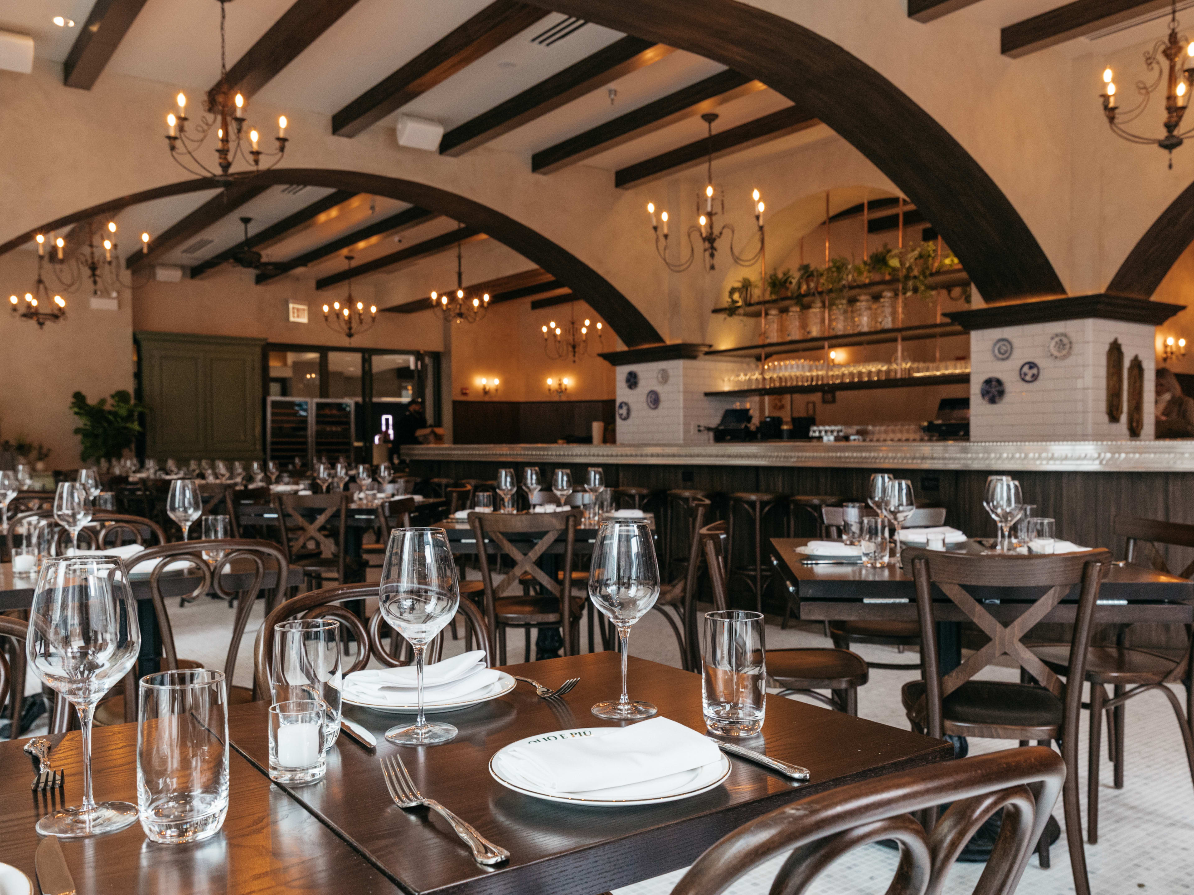 The interior of Olio e Piu. There are vaulted ceilings, wooden tables set with dishware and glasses and a wrap around bar.