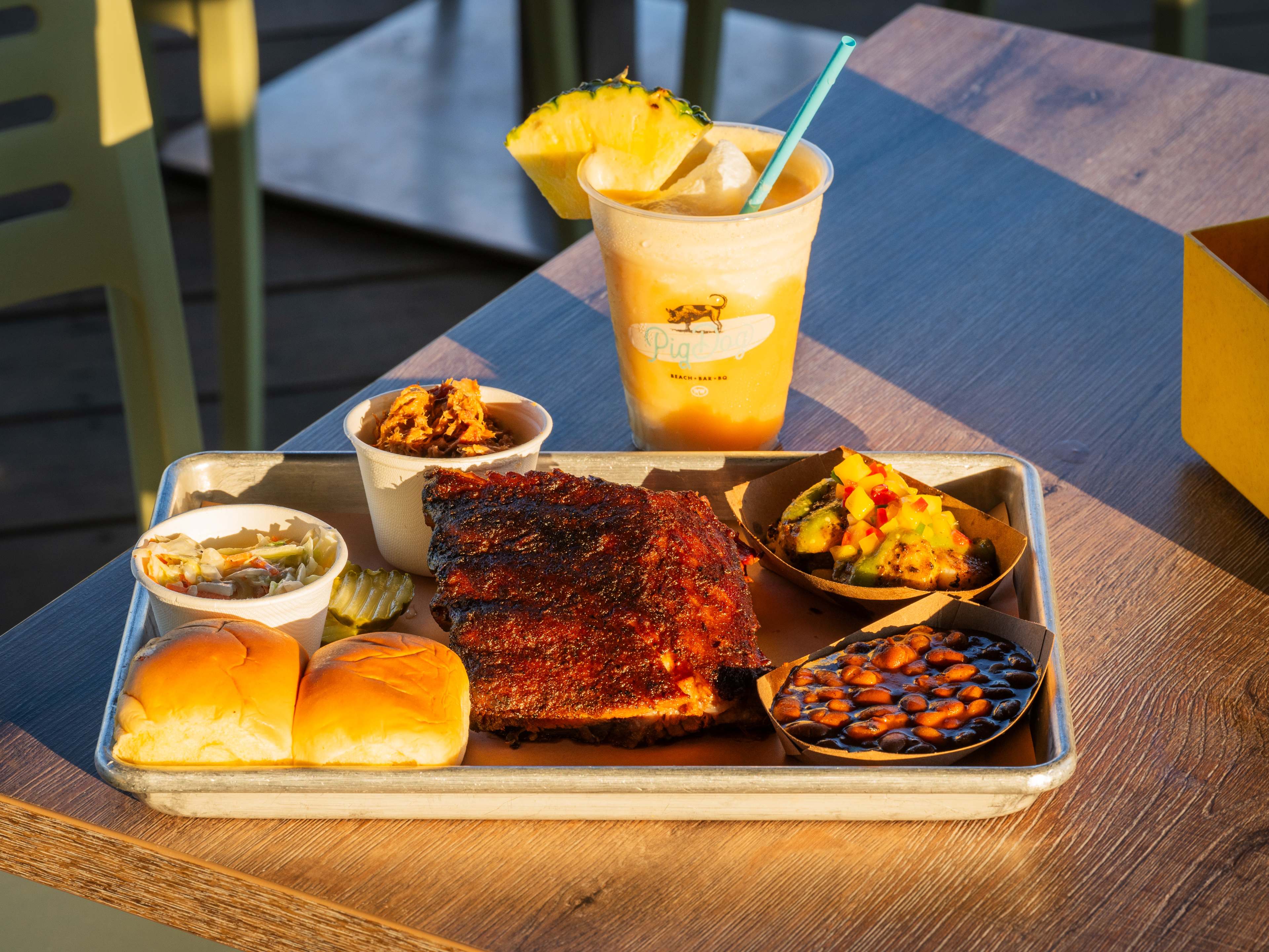 Tray of food containing the Cuban Scallops and BBQ Combo accompanied by a Piña Colada on a wooden table.