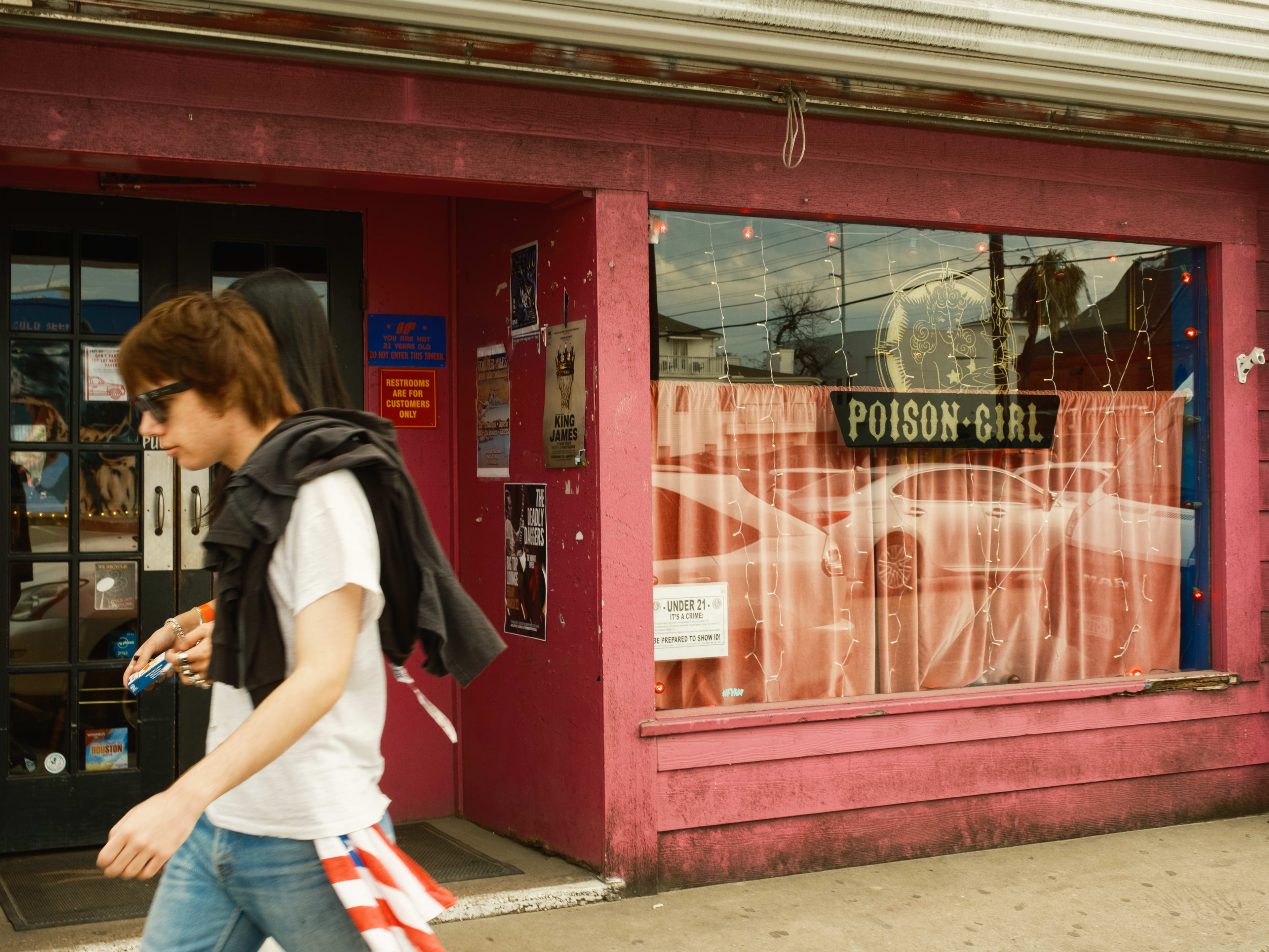 The red exterior of Poison Girl. A person with a white t-shirt and sunglasses walks by.