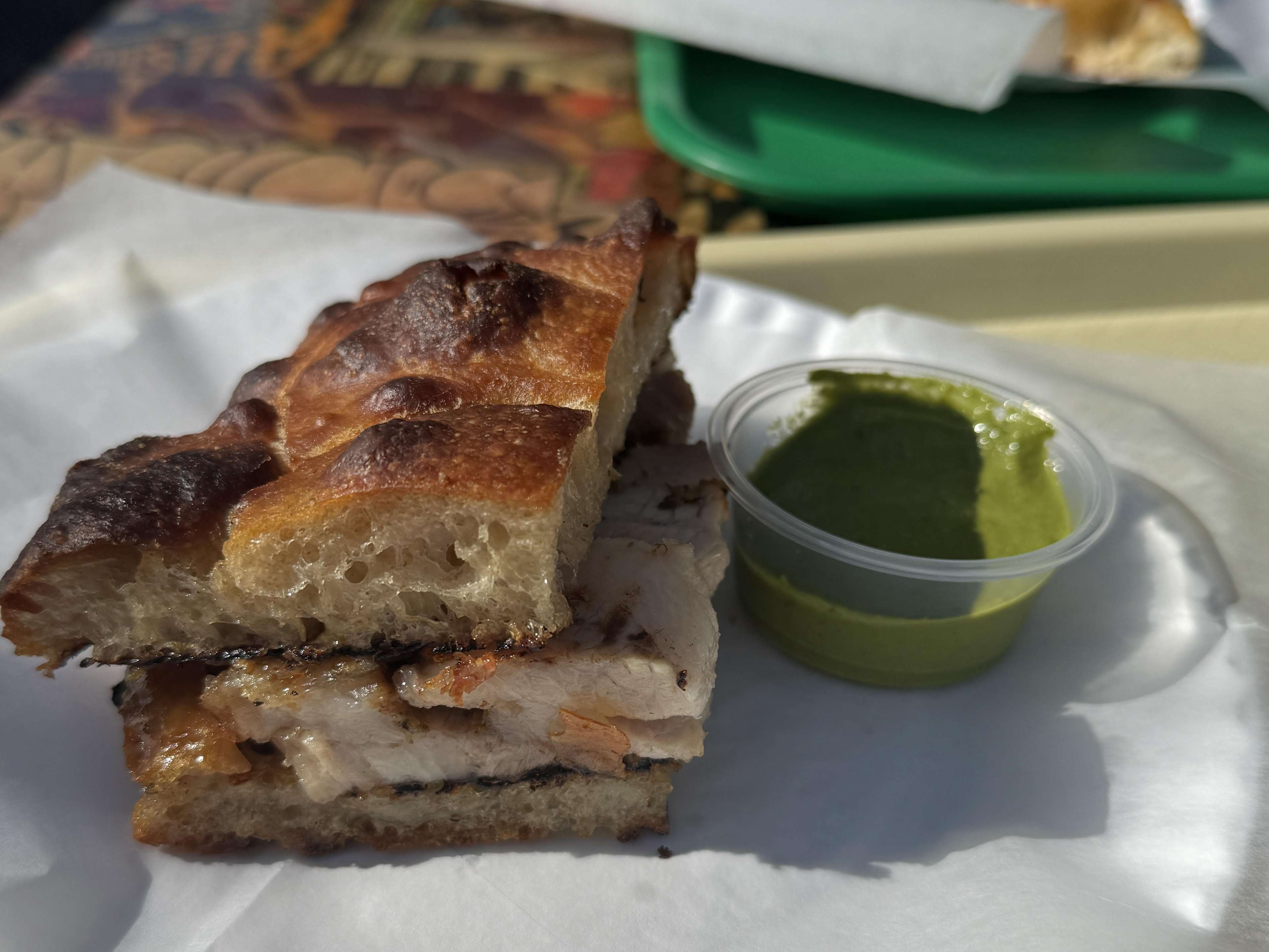 A porchetta sandwich and small container of salsa verde on a paper plate.