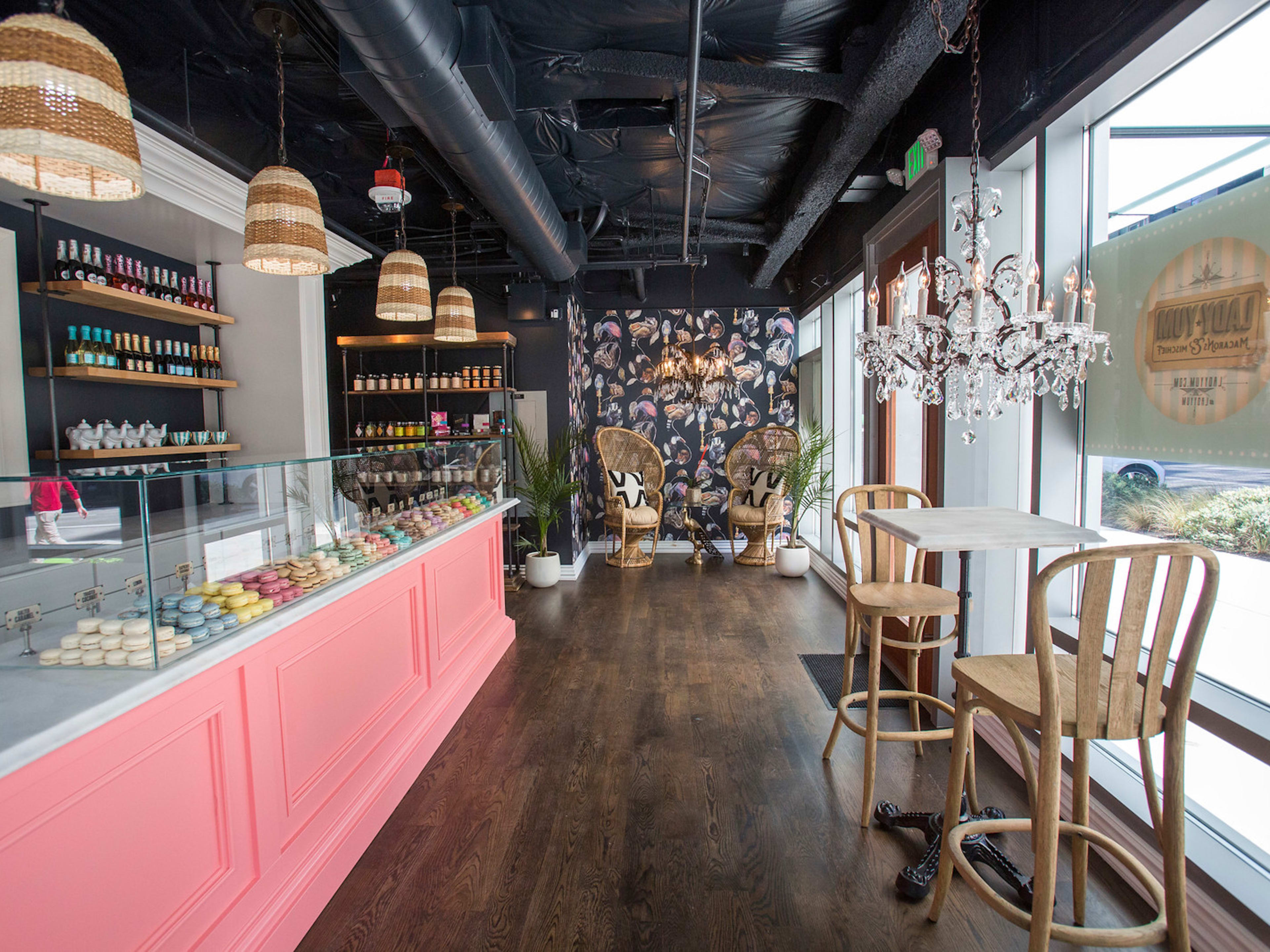 A macaron bakery interior with shelves of miniature sparkling wine bottles, macarons, and a couple of wicker chairs.