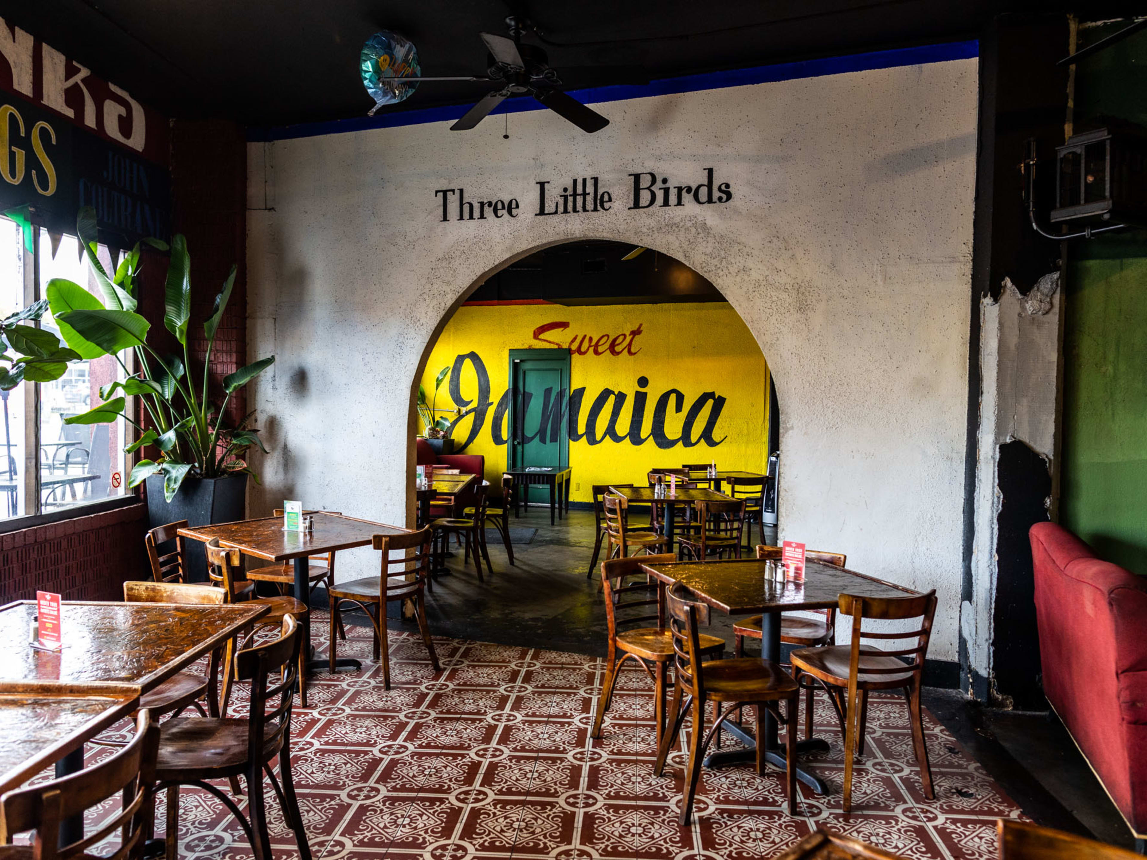 The interior of Reggae Hut with a painted mural reading, "Sweet Jamaica" and "Three Little Birds" painted above an archway. There is patterned laminate flooring, wooden tables and chairs.