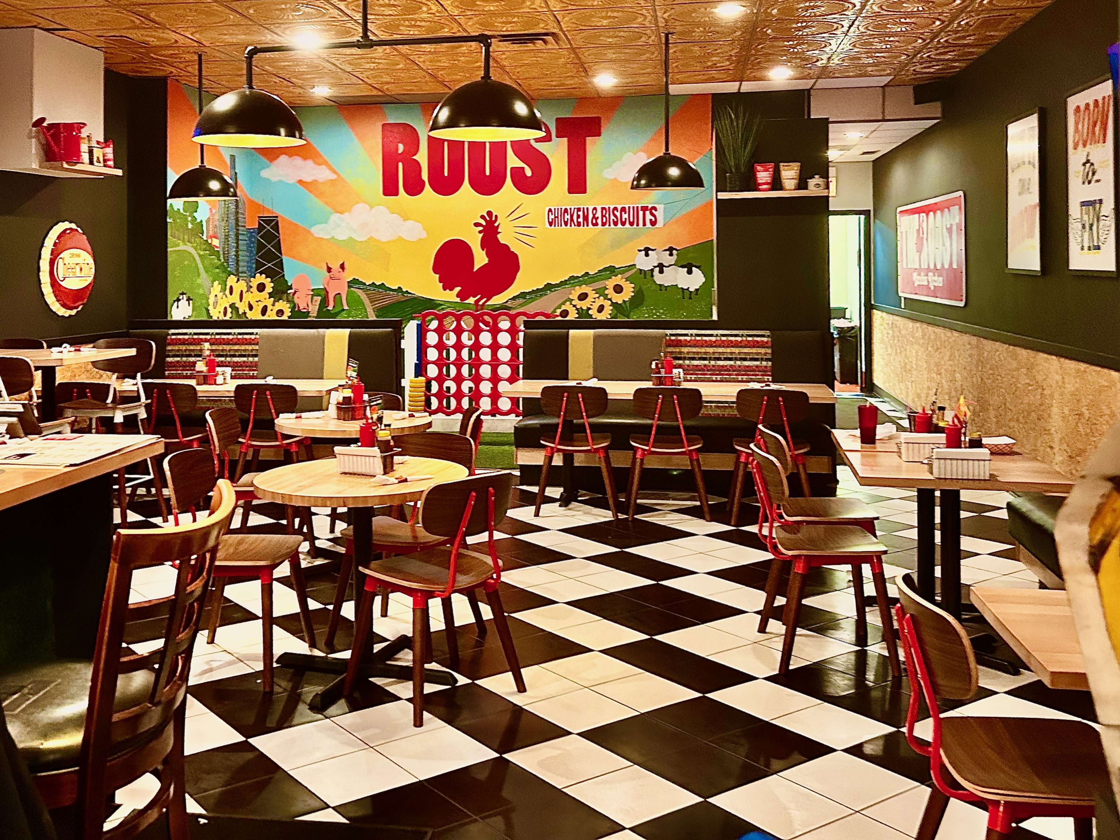 A colorful retro interior with a checkerboard tile  floor, empty tables, and a giant Roost Chicken & Biscuits mural in the background.