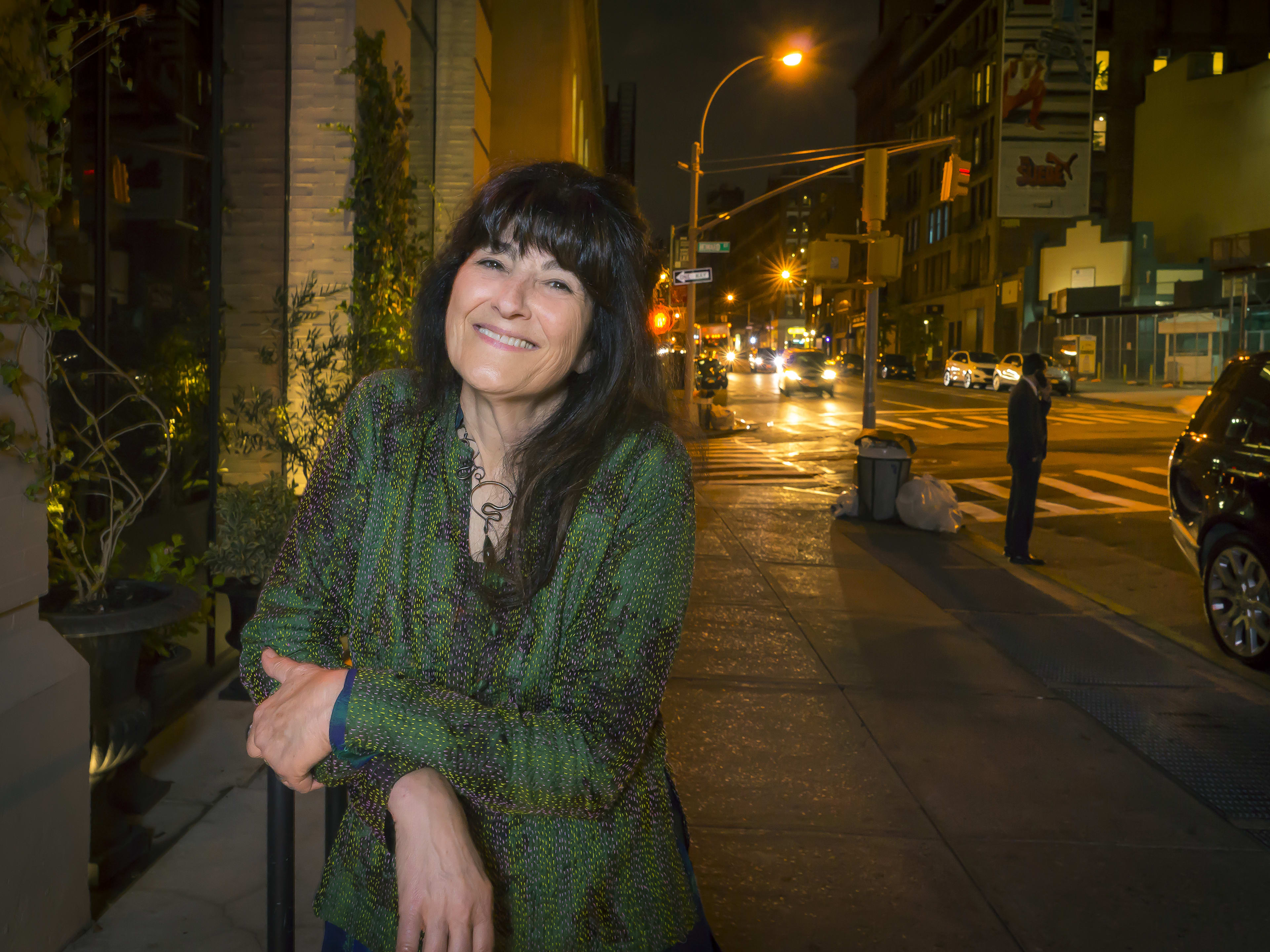 the food writer Ruth Reichl stands with her arms crossed, wearing a green outfit, on a dark nighttime New York City sidewalk as cars approach on the street behind her