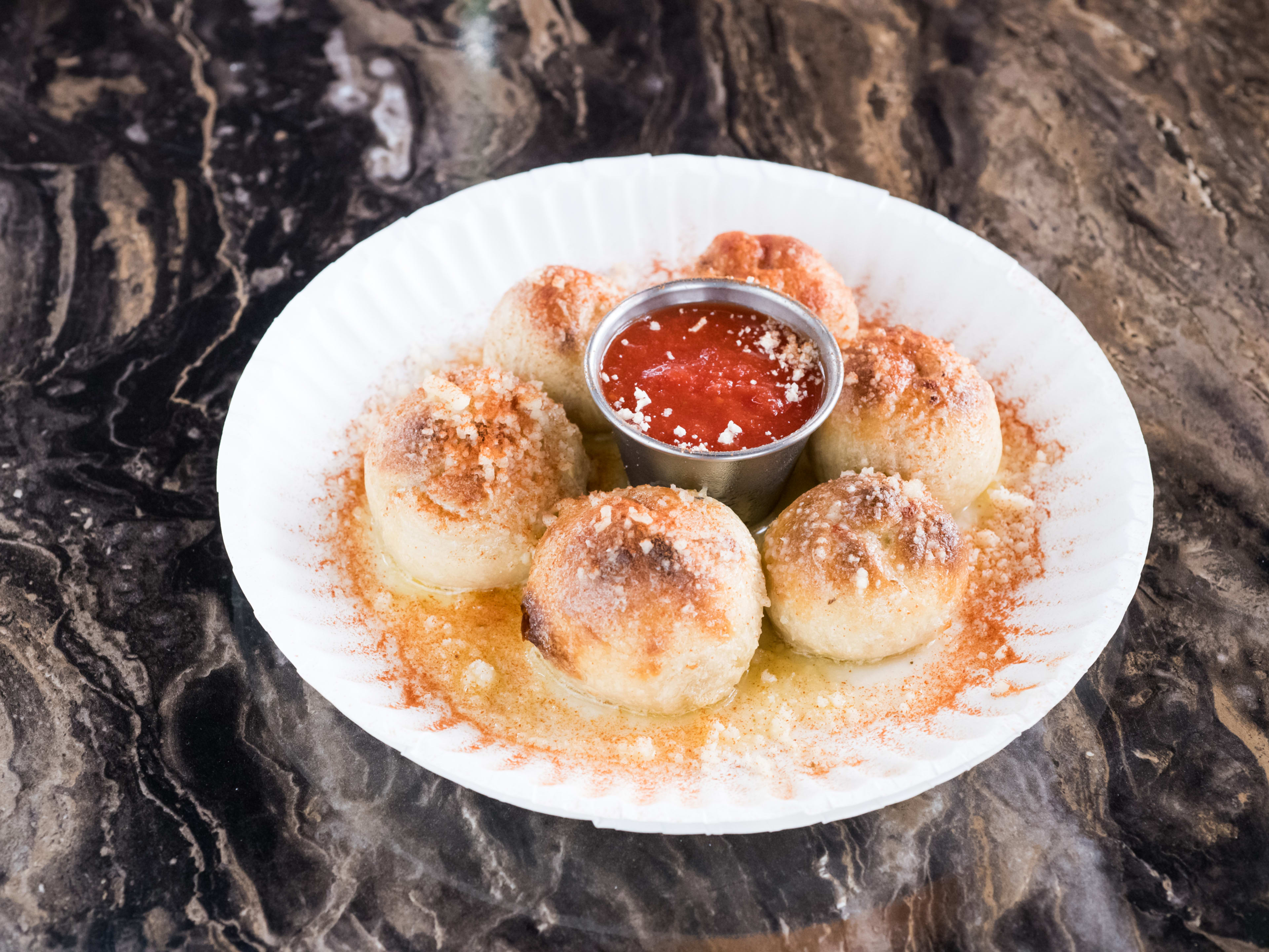 A marble table with a paper plate of garlic knots and a ramekin of marinara sauce