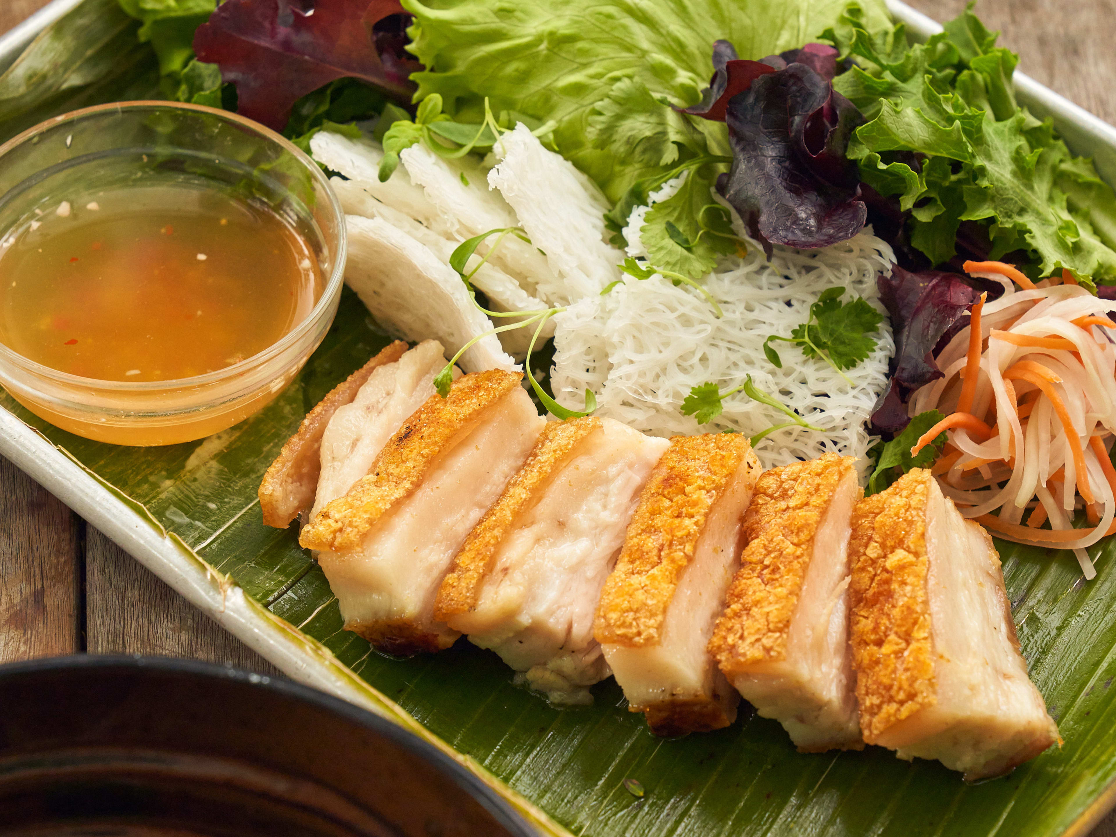 Leaf lined tray with sliced pork belly, lettuce, carrots, noodles, and dipping sauce.