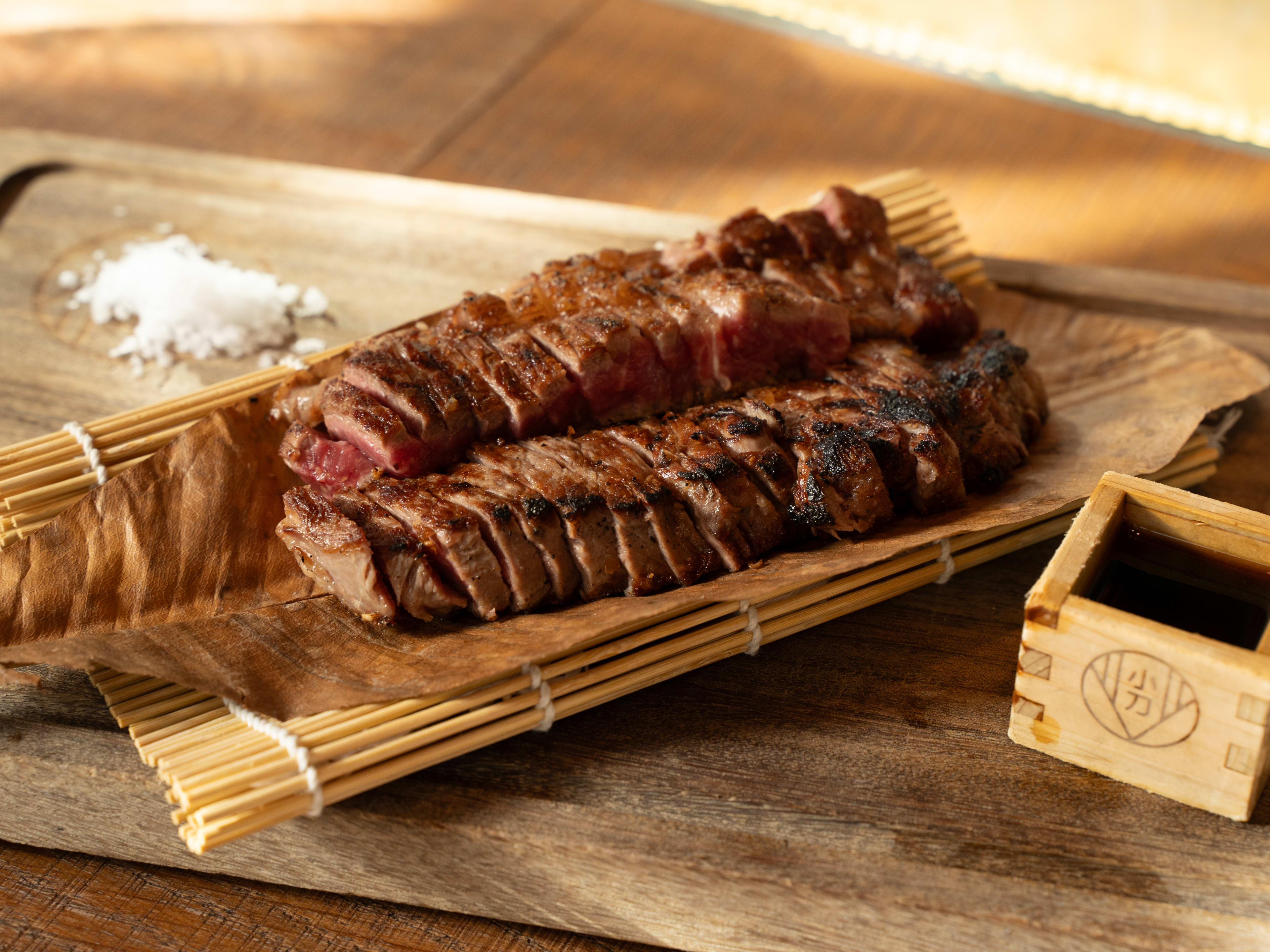 Massive wagyu steak served on bamboo mat next to pile of flaky salt and wooden container of sauce.