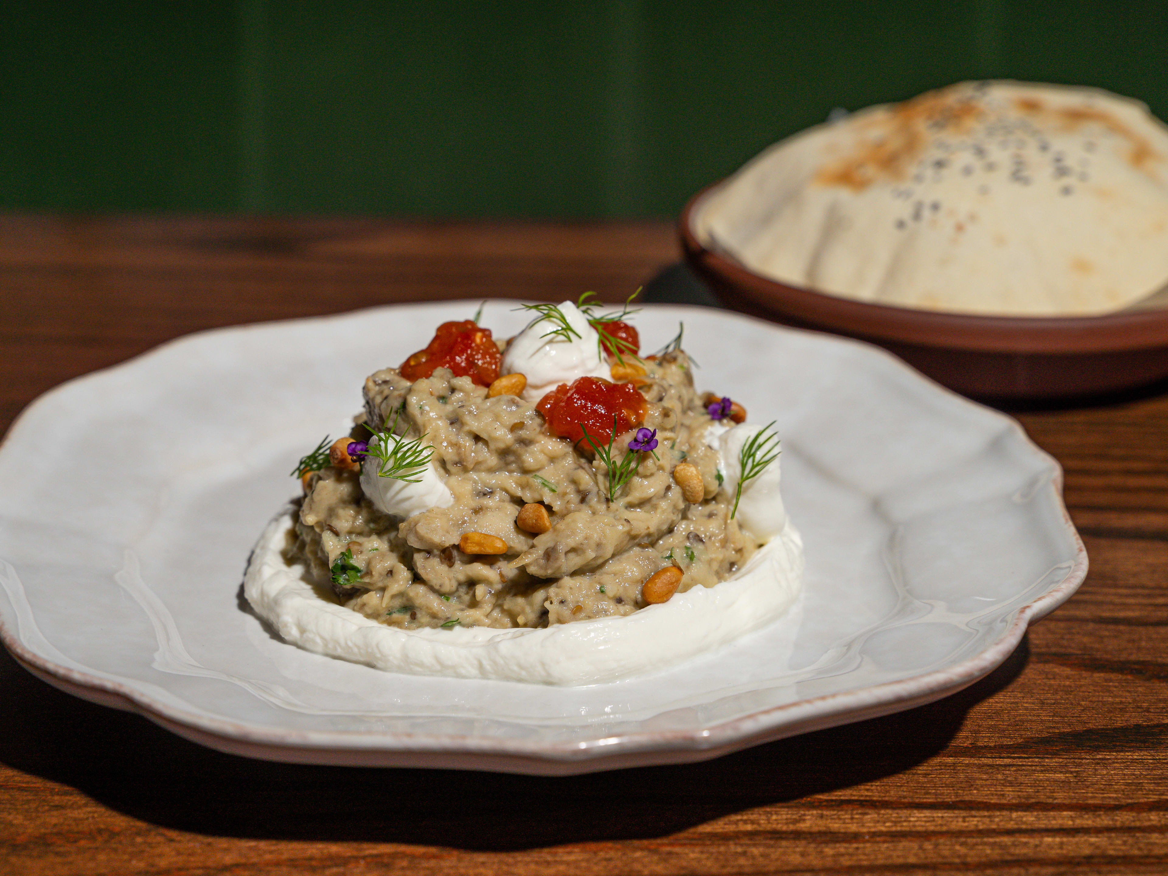 Mound of mashed eggplant topped with confit tomatoes and pine nuts on a layer of labneh with pita on the side