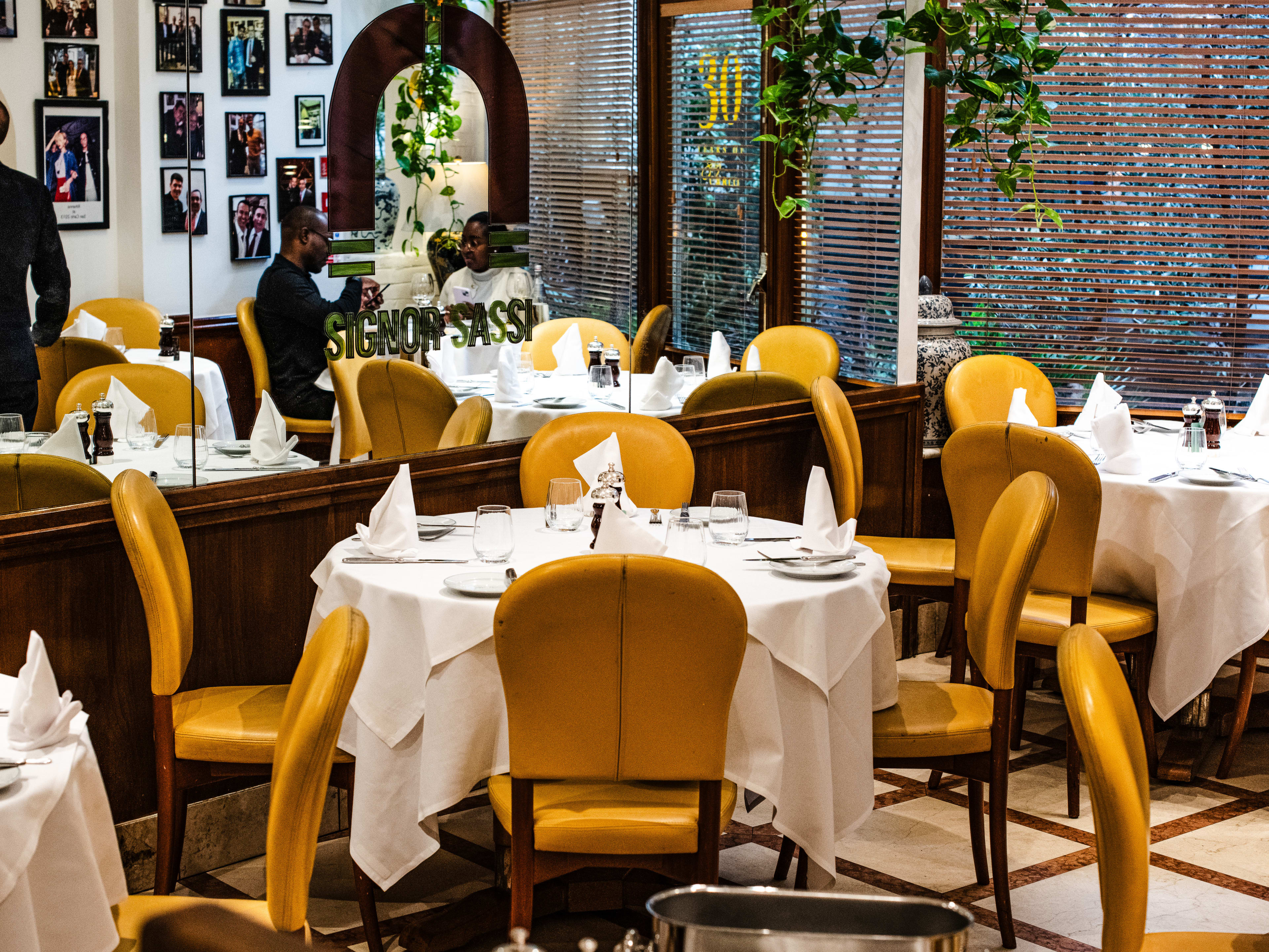 The interior of Signor Sassi. There are small round tables with white tablecloths and yellow chairs. There are two people dining at a table, reflected in the mirrored-wall. Also reflected is a gallery wall of celebrity headshots.