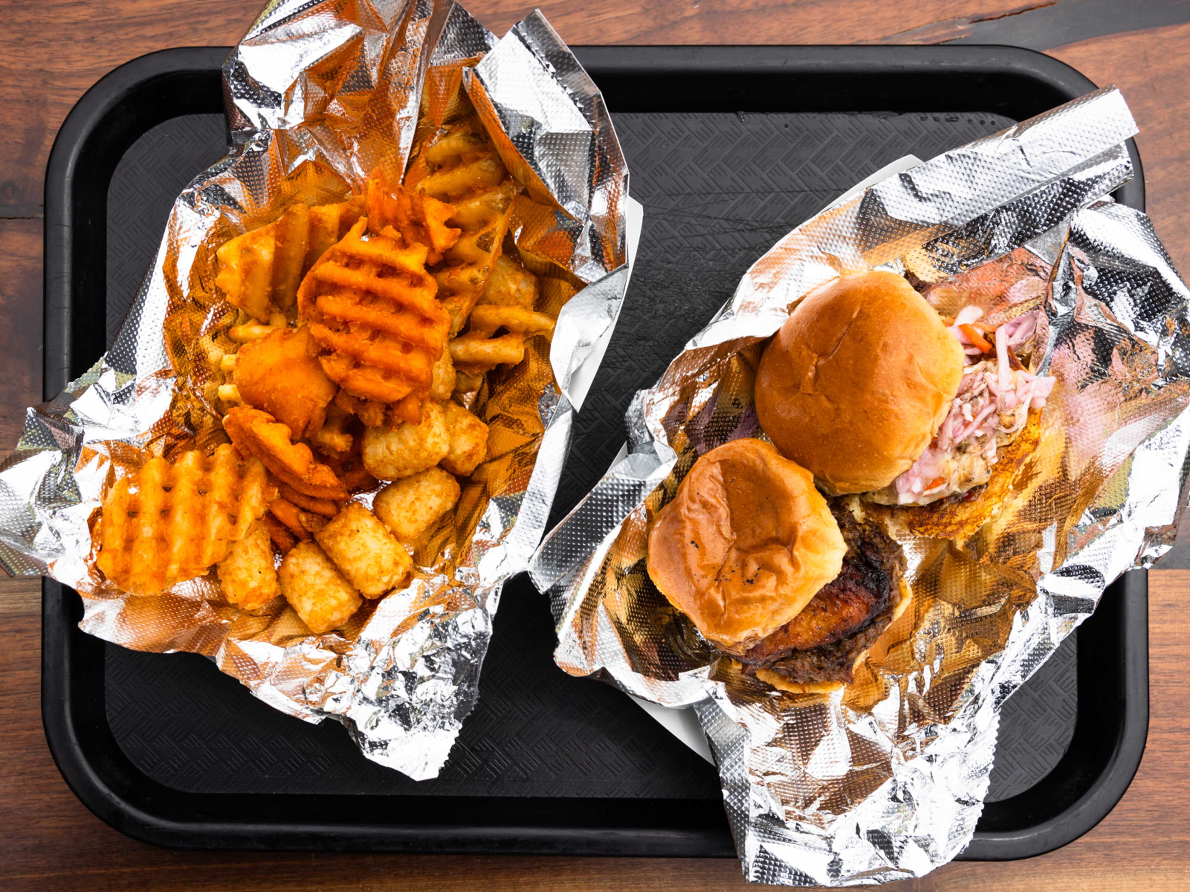 Two sliders and an assortment of french fries and tater tots on a black tray.