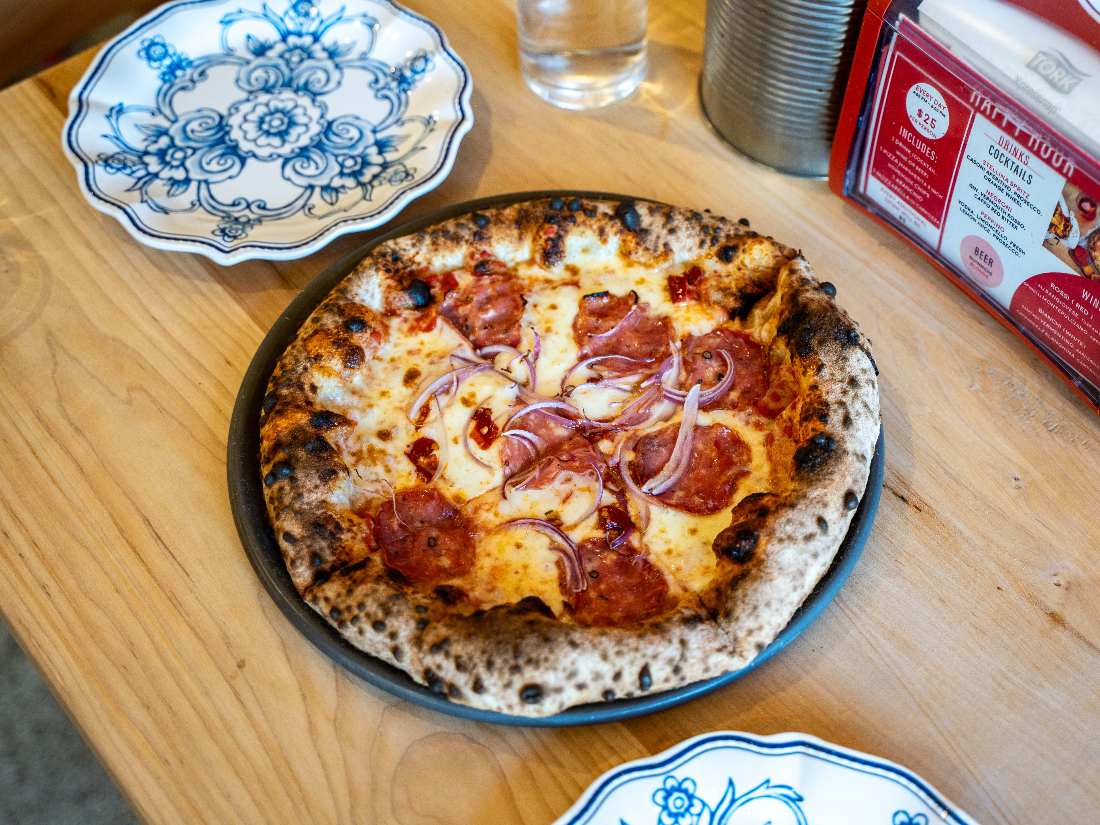 Pepperoni pizza with charred crust and red onions on table with two china plates.