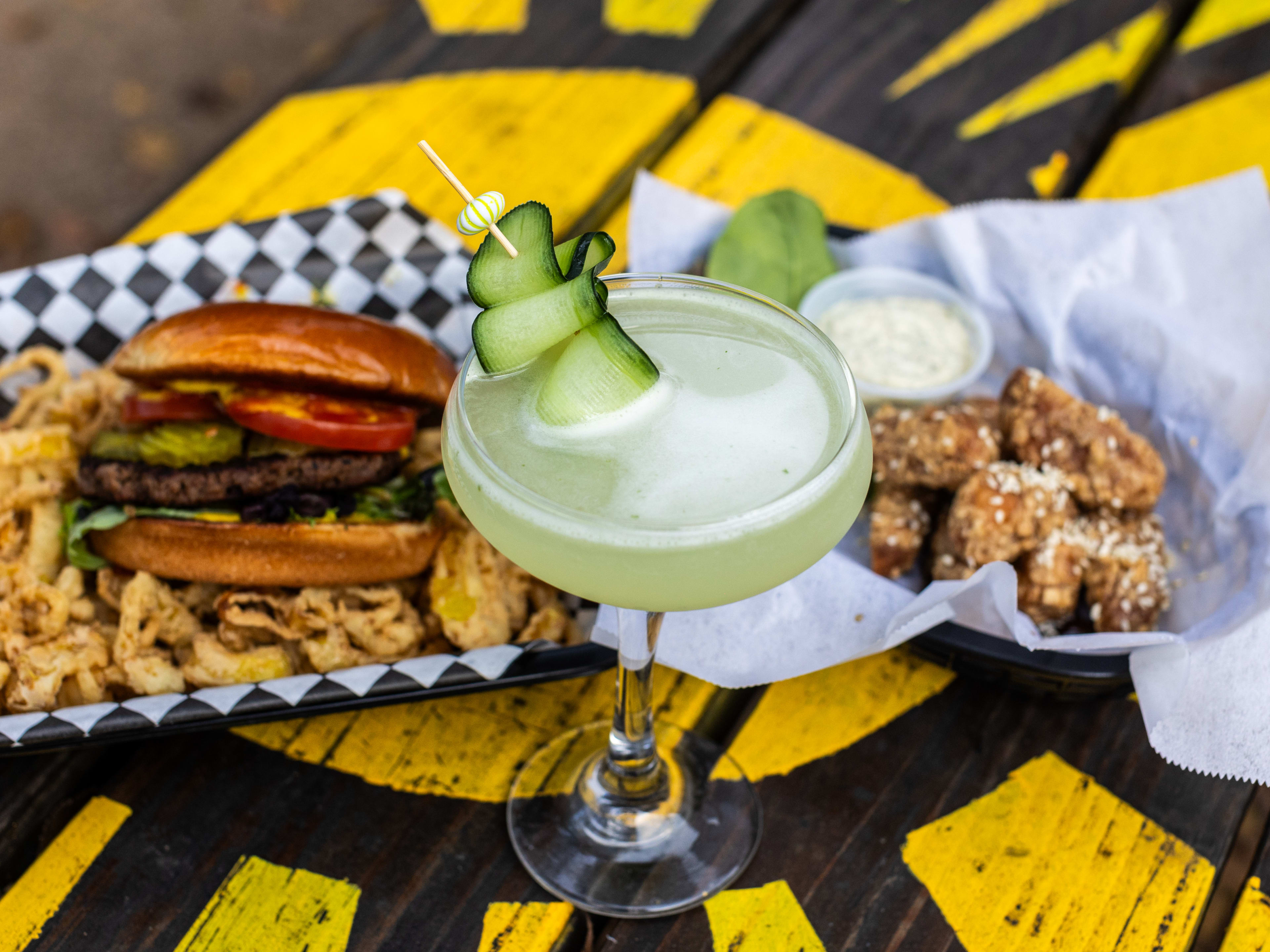 Baskets with a burger and fried banana peppers, and wings, with a cucumber garnished cocktail on a yellow and black picnic table.