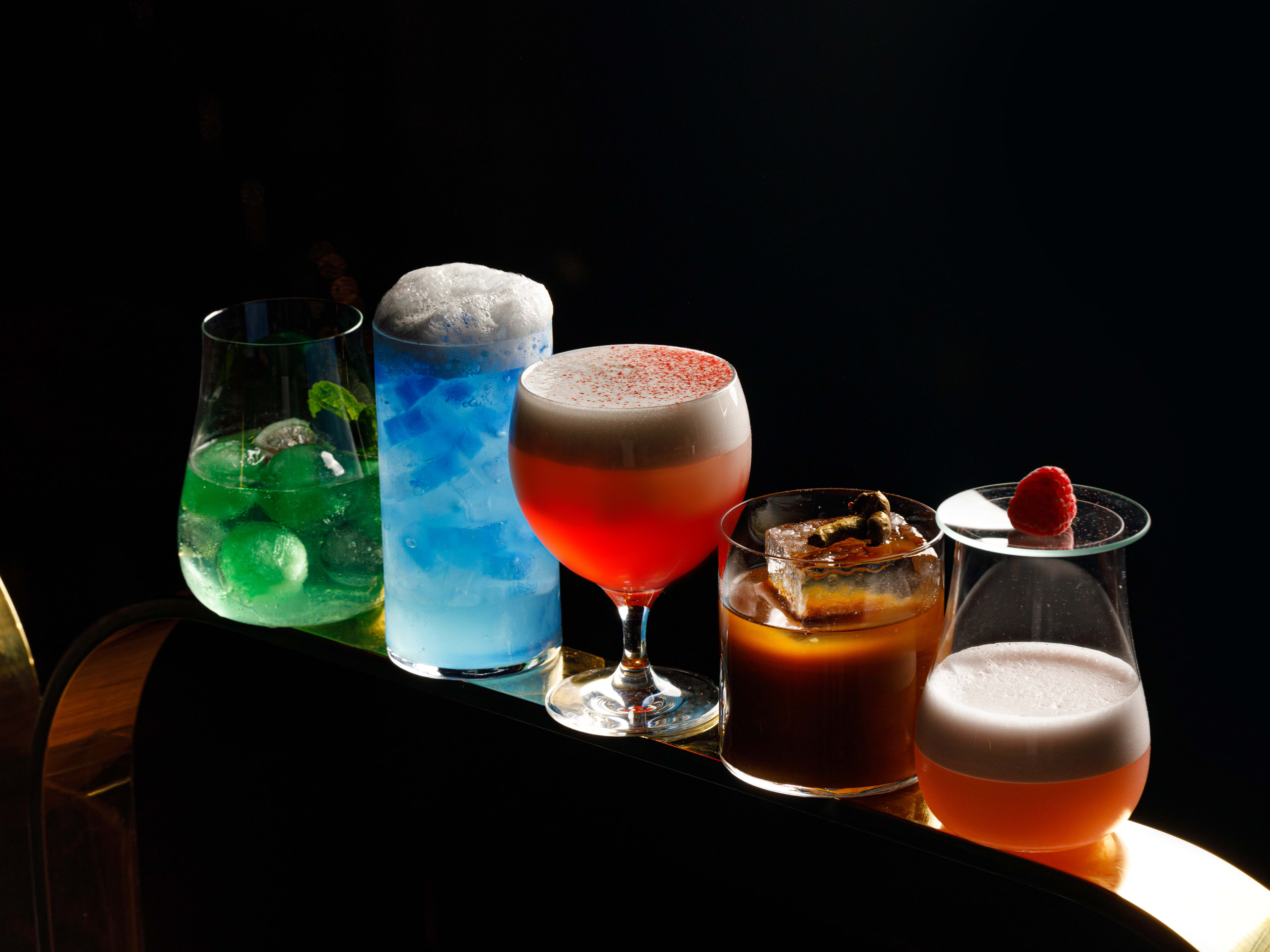 Five colorful cocktails lined up in different fancy glasses