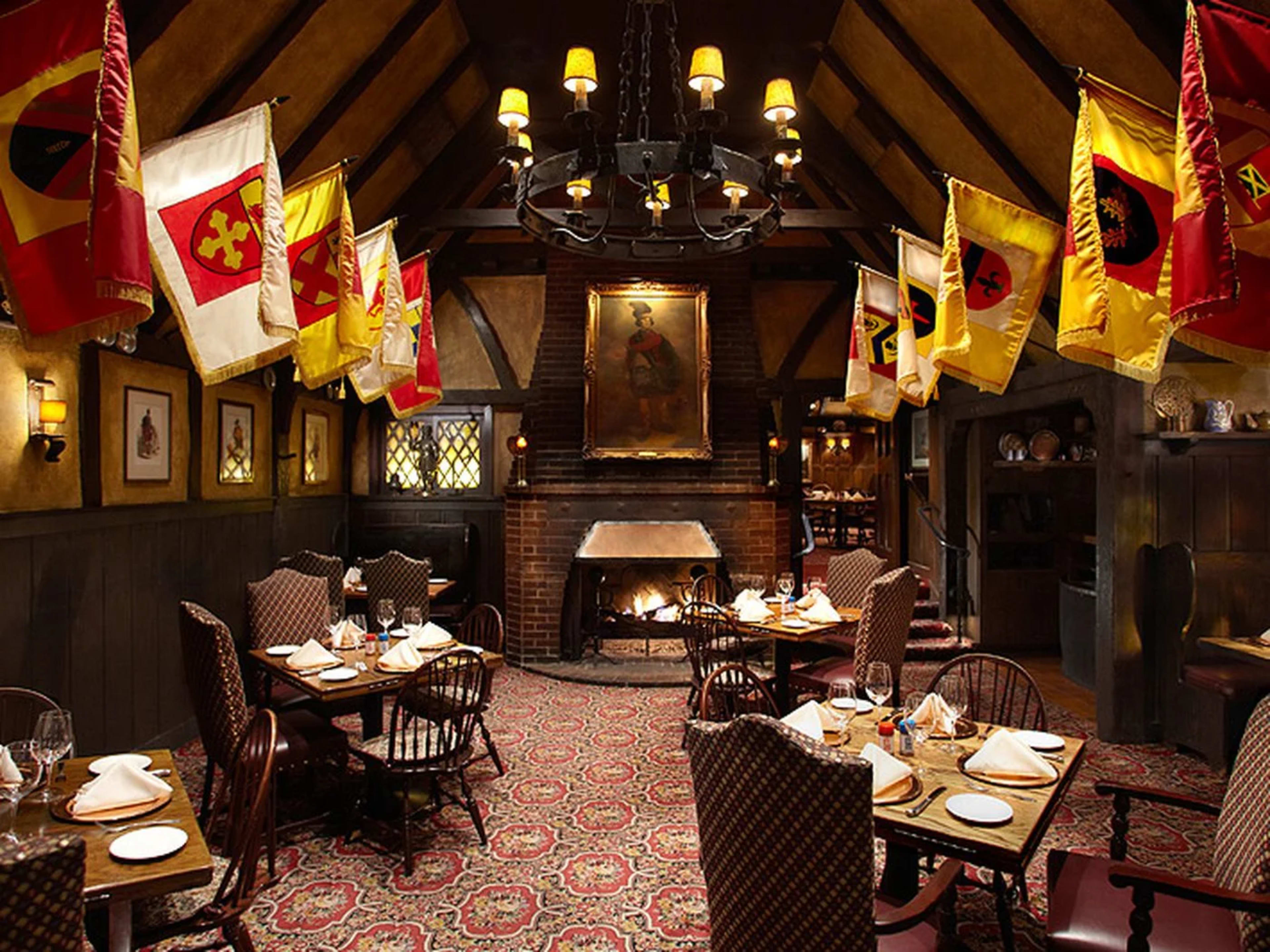 The mediaeval style interior of Tam O’ Shanter. There are crest flags hanging on the walls, a chandelier, and a portrait hanging over a lit fireplace. There are wooden table with patterned fabric covered chairs.