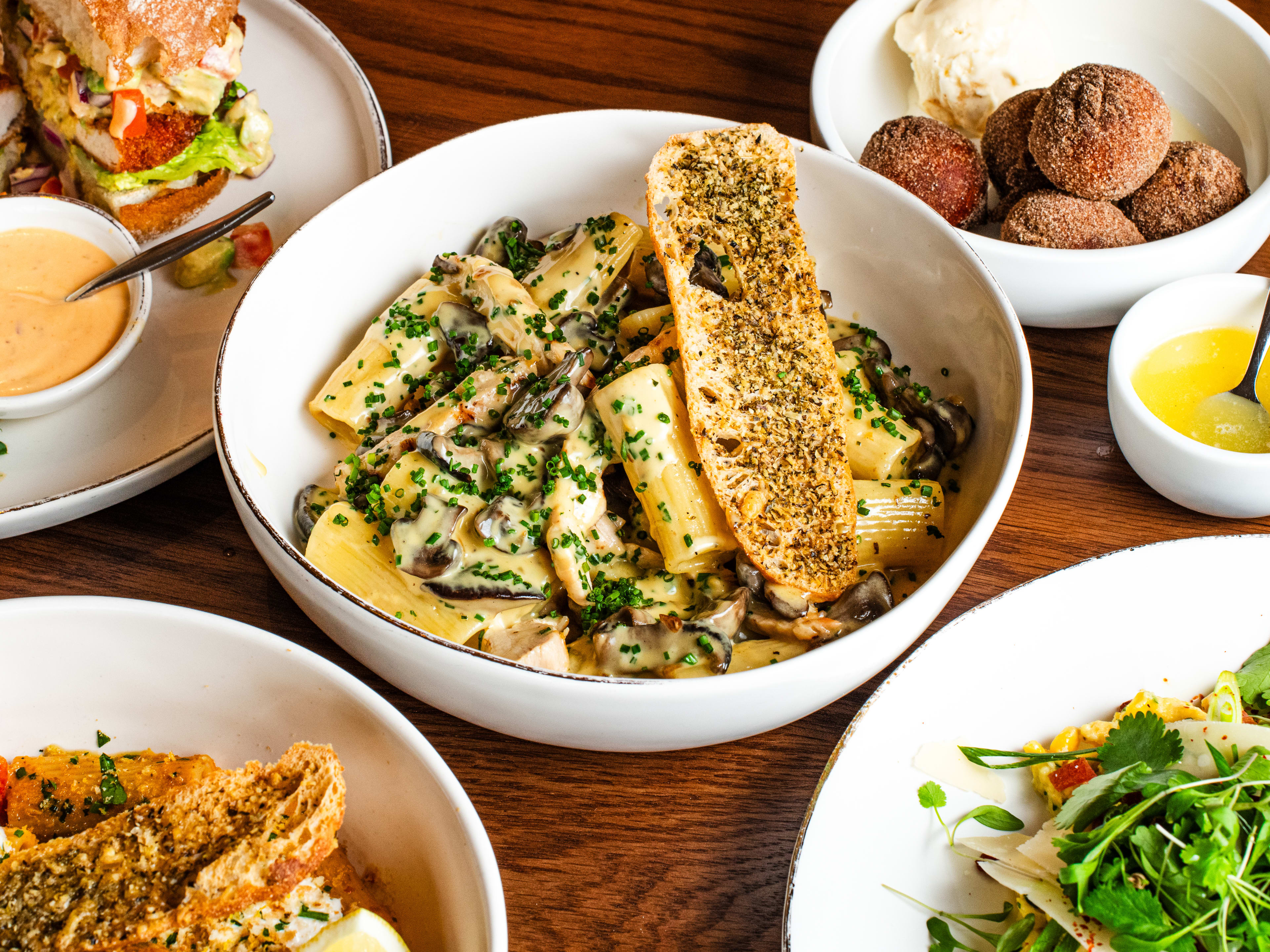 Various dishes from Tashas sit on a wooden table. Savva's Chicken Pasta is in the center.