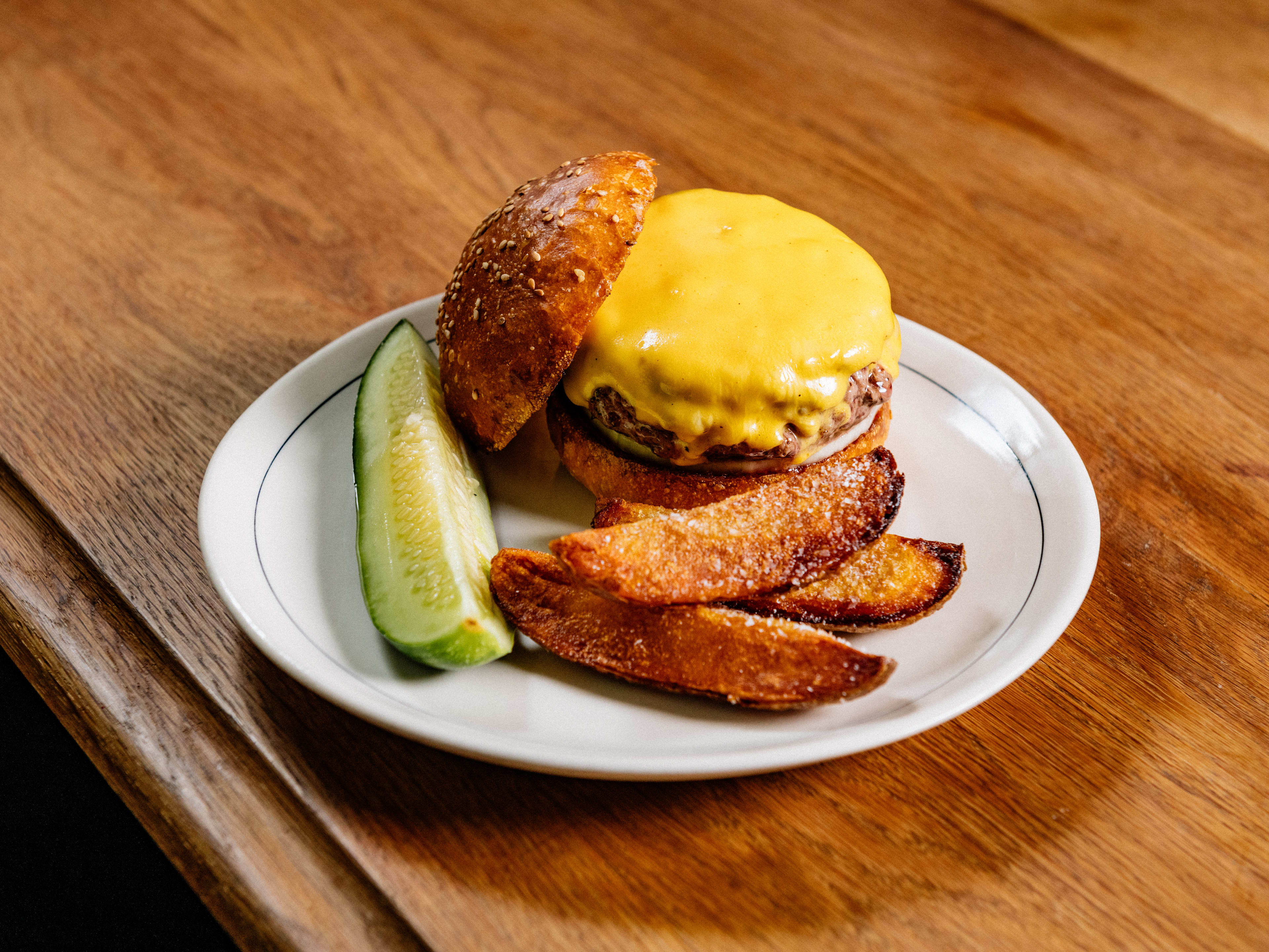 A cheeseburger burger, potato wedges and a pickle spear on a white plate sitting on a wooden bar.