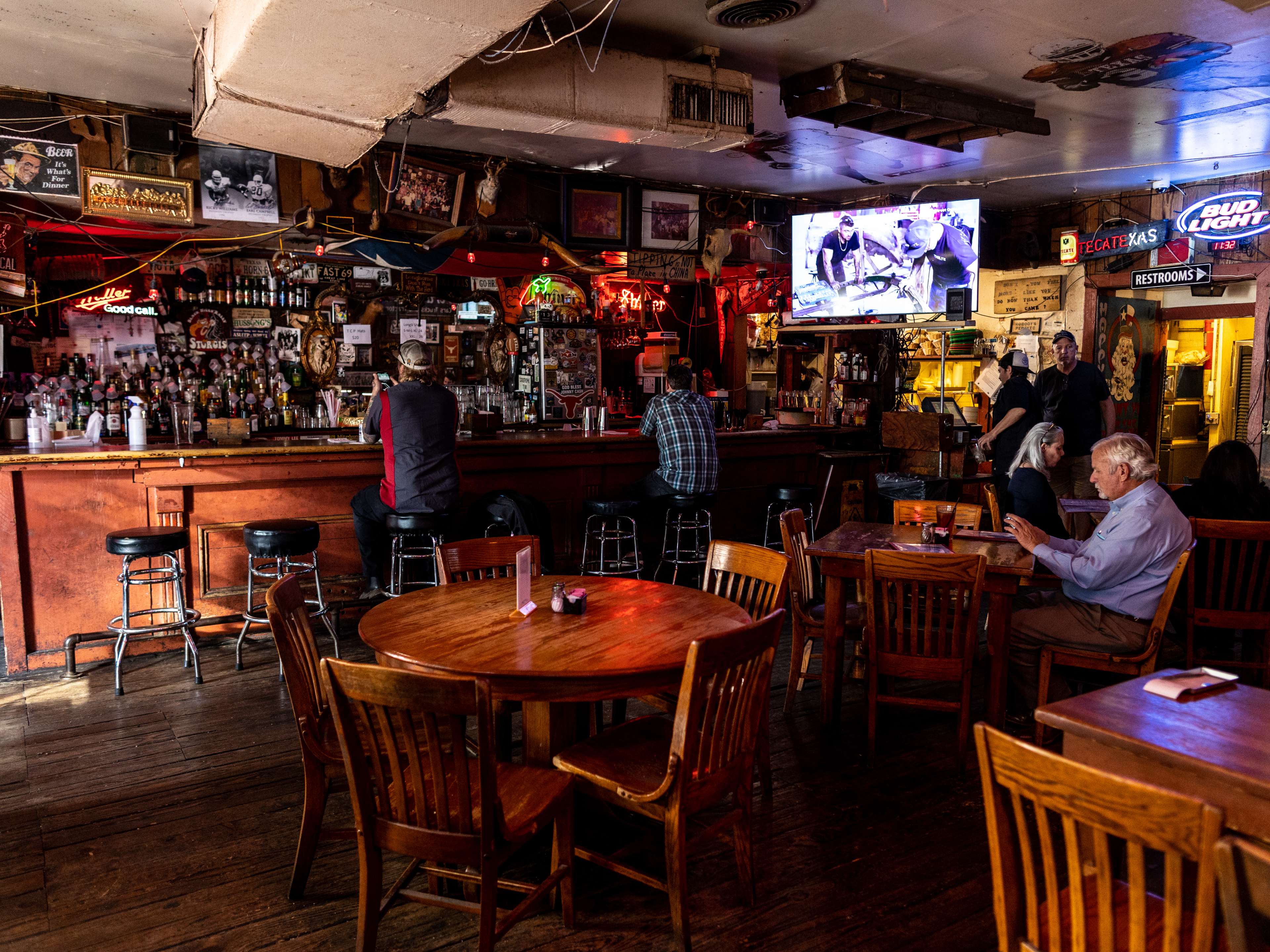 The interior of Texas Chili Parlor. There are people sitting at a wooden bar, and wooden tables and chairs.  A TV hung above the bar is playing sports.