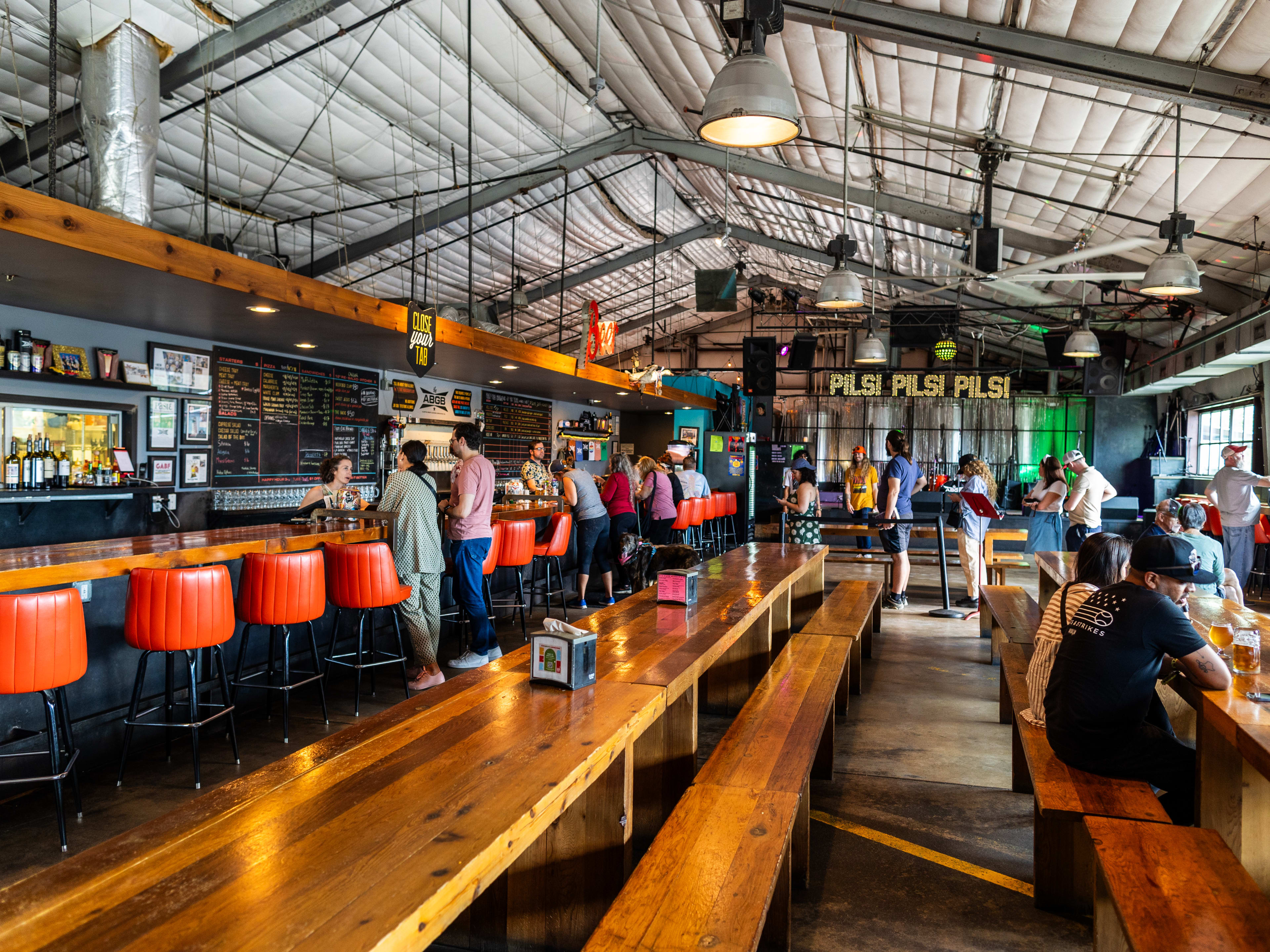 The interior of The ABGB with long communal tables, a long bar with orange leather seats and industrial fans and lights hanging from the metal garage ceiling.