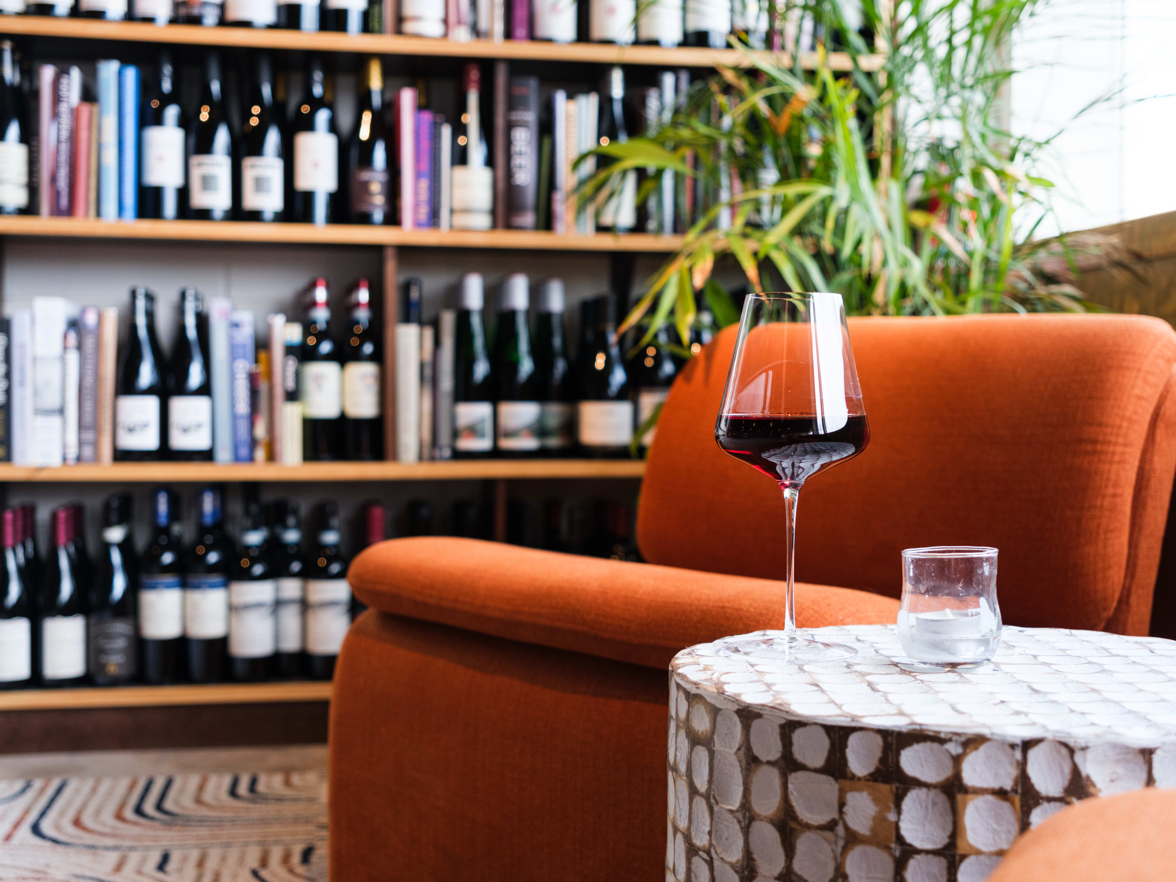 A glass of red wine on a round side table next to a mod orange lounge chair in front of a plant and a bookshelf mixed with wine bottles and books.