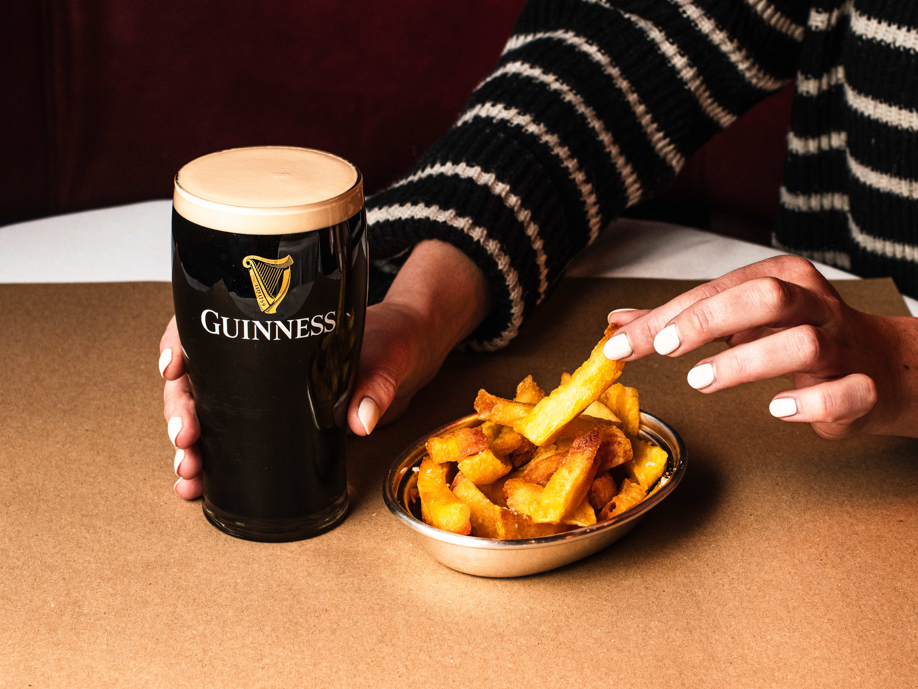 A woman's hand reaching for a chip with one hand and holding a pint of Guinness in the other.