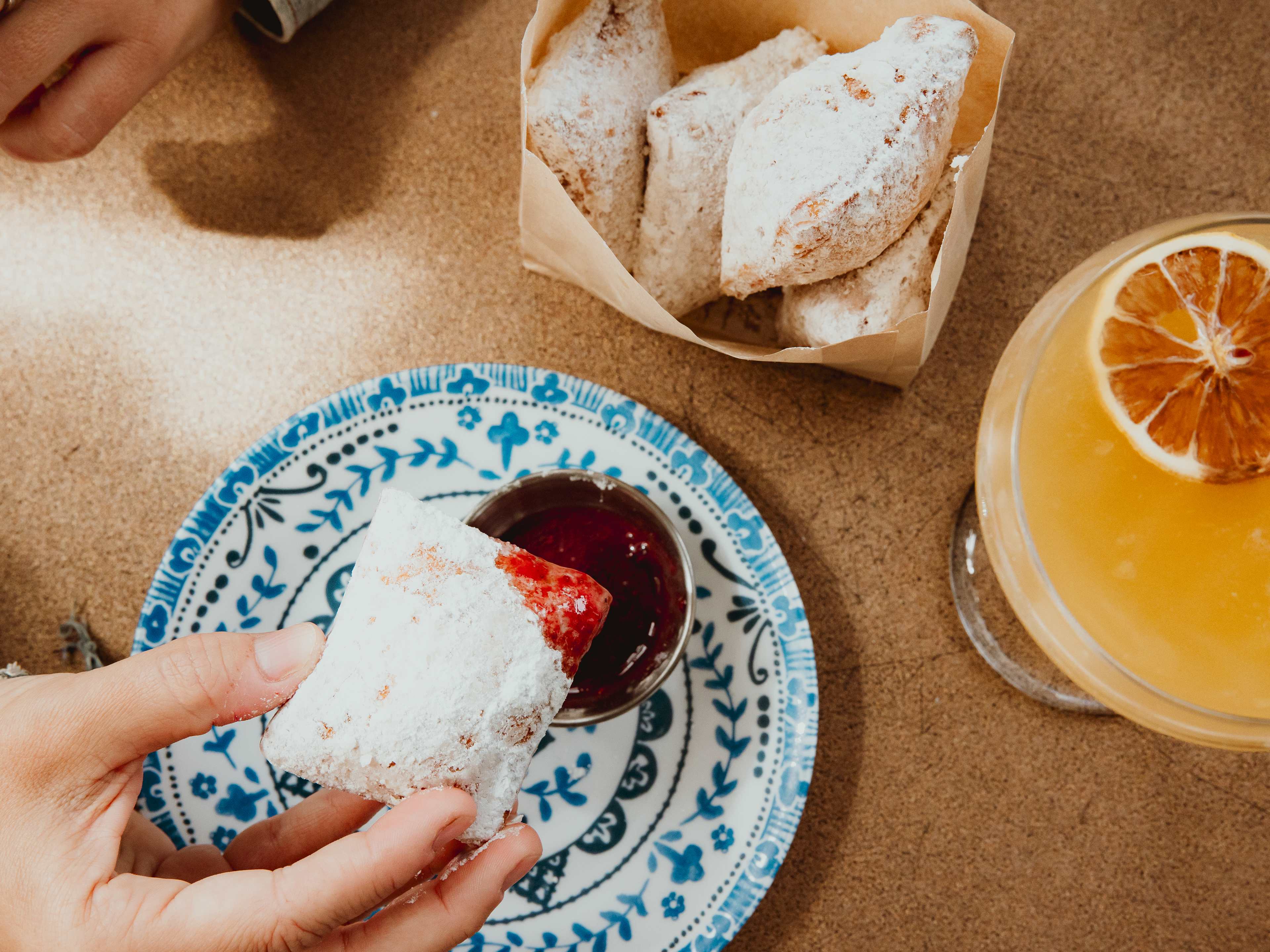 bag of beignets next to cocktail, beignet being dipped into raspberry sauce