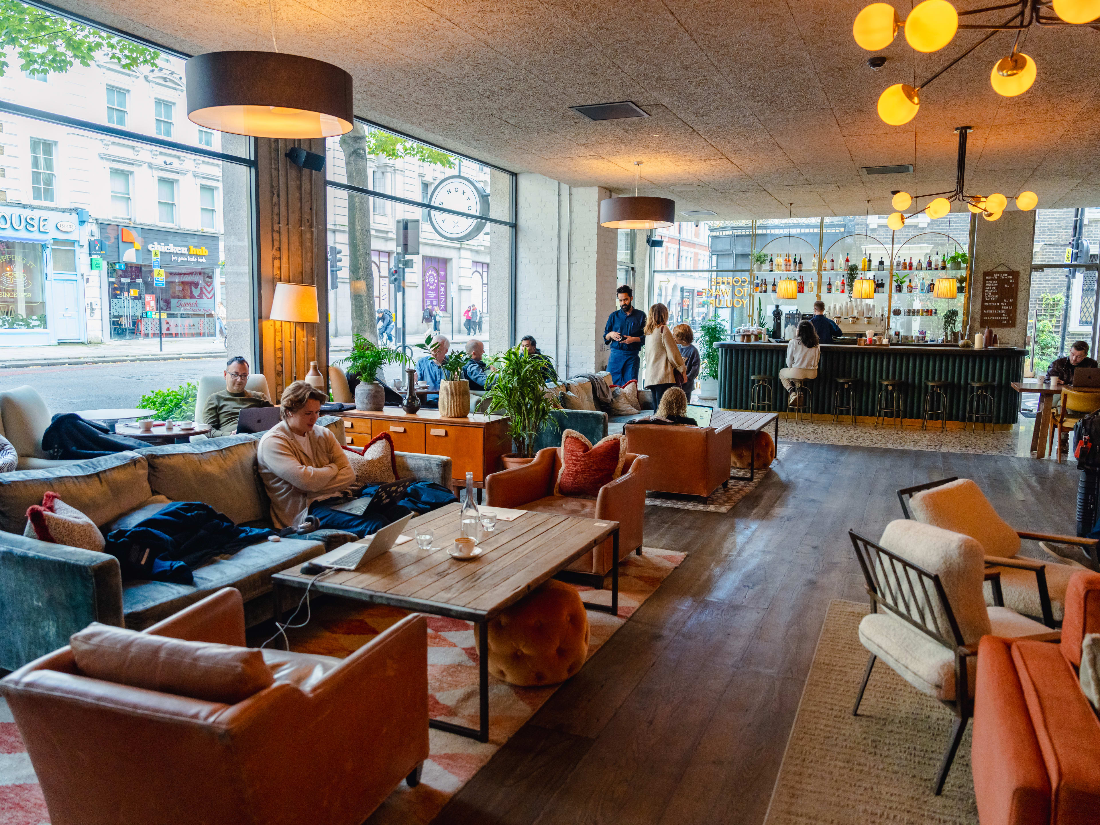 The interior of The Hoxton with pink and blue velvet couches and chairs, people sitting with laptops, or standing and chatting. There is a green bar at the back with an espresso machine.