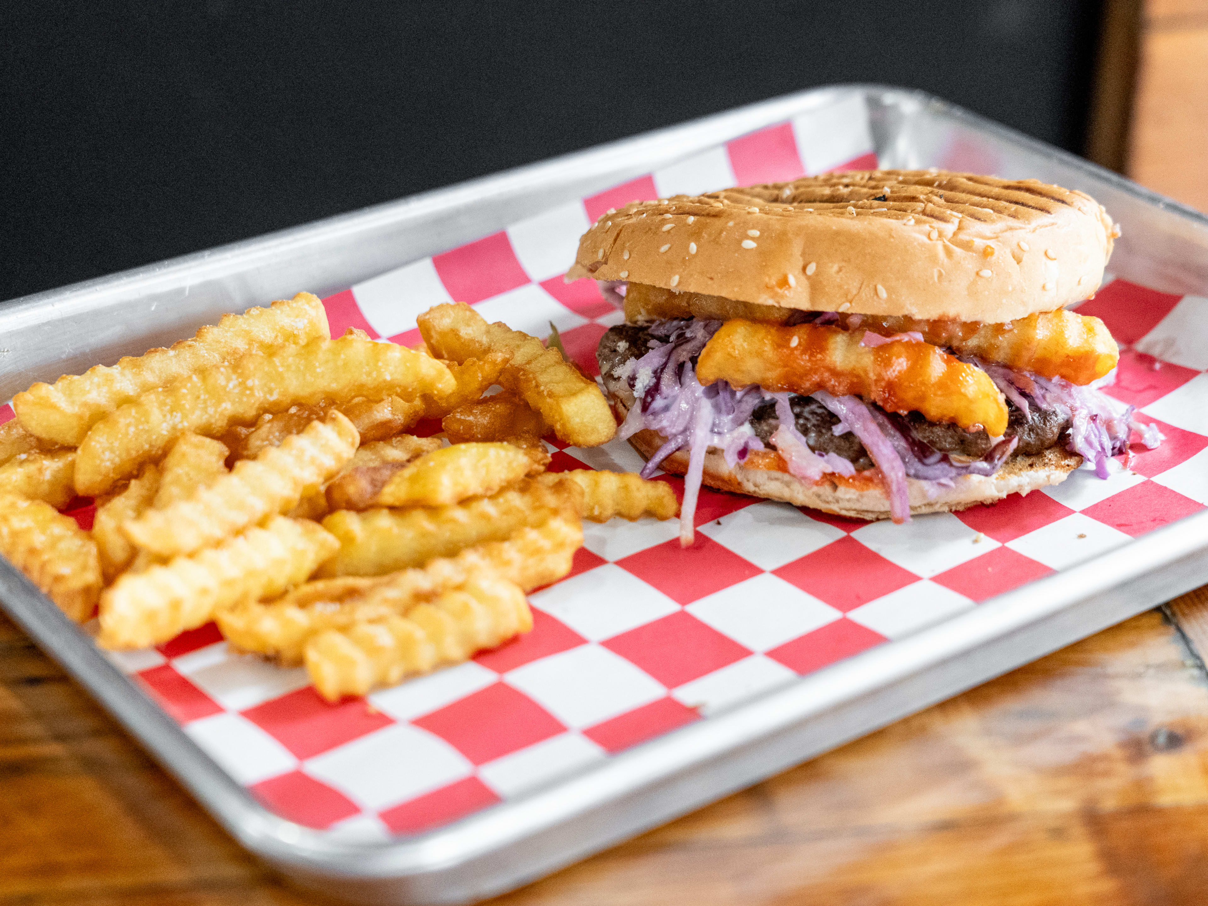 Burger on seeded bun filled with red onion and fries, next to crinkle cut fries on paper-lined tray.