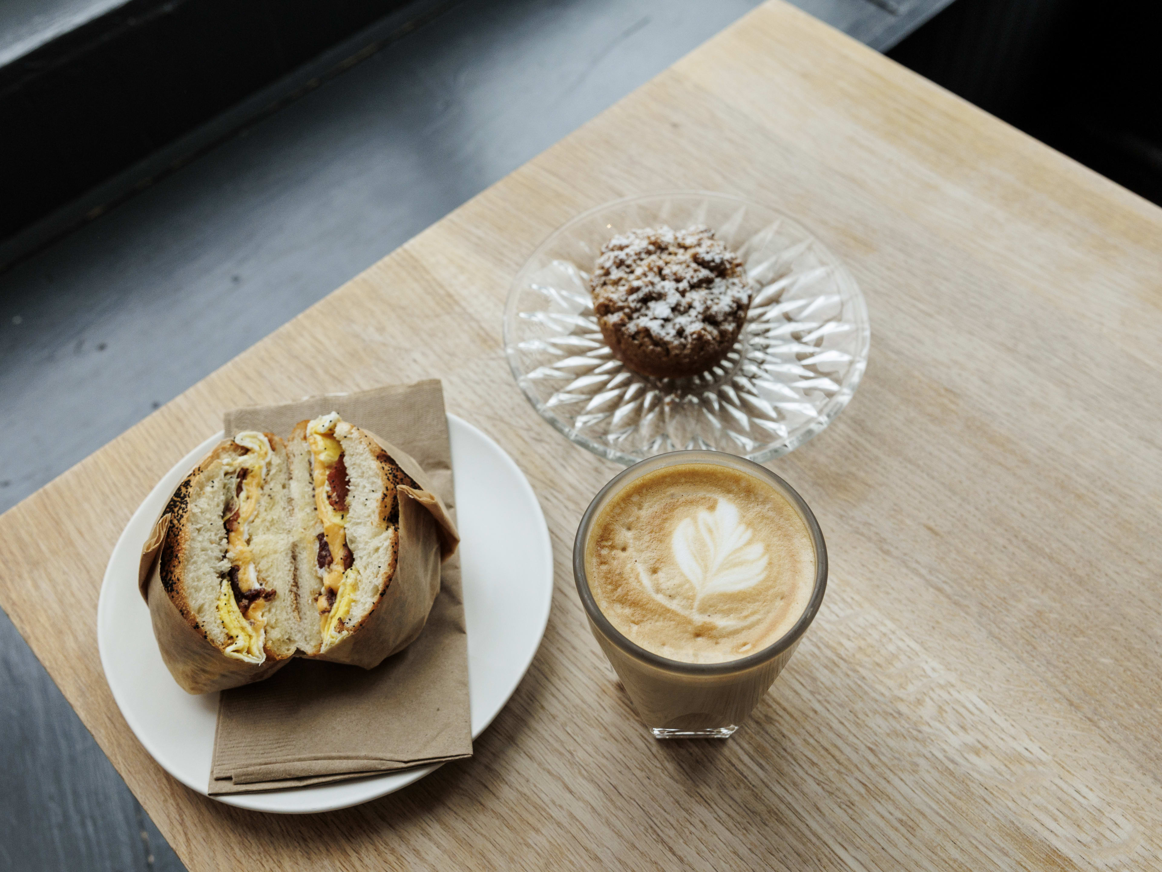 A wooden table with bacon, egg, and cheese on a poppy seed kaiser roll, a cortado with latte art, and a crystal plate holding a miniature powdered sugar-covered crumb cake