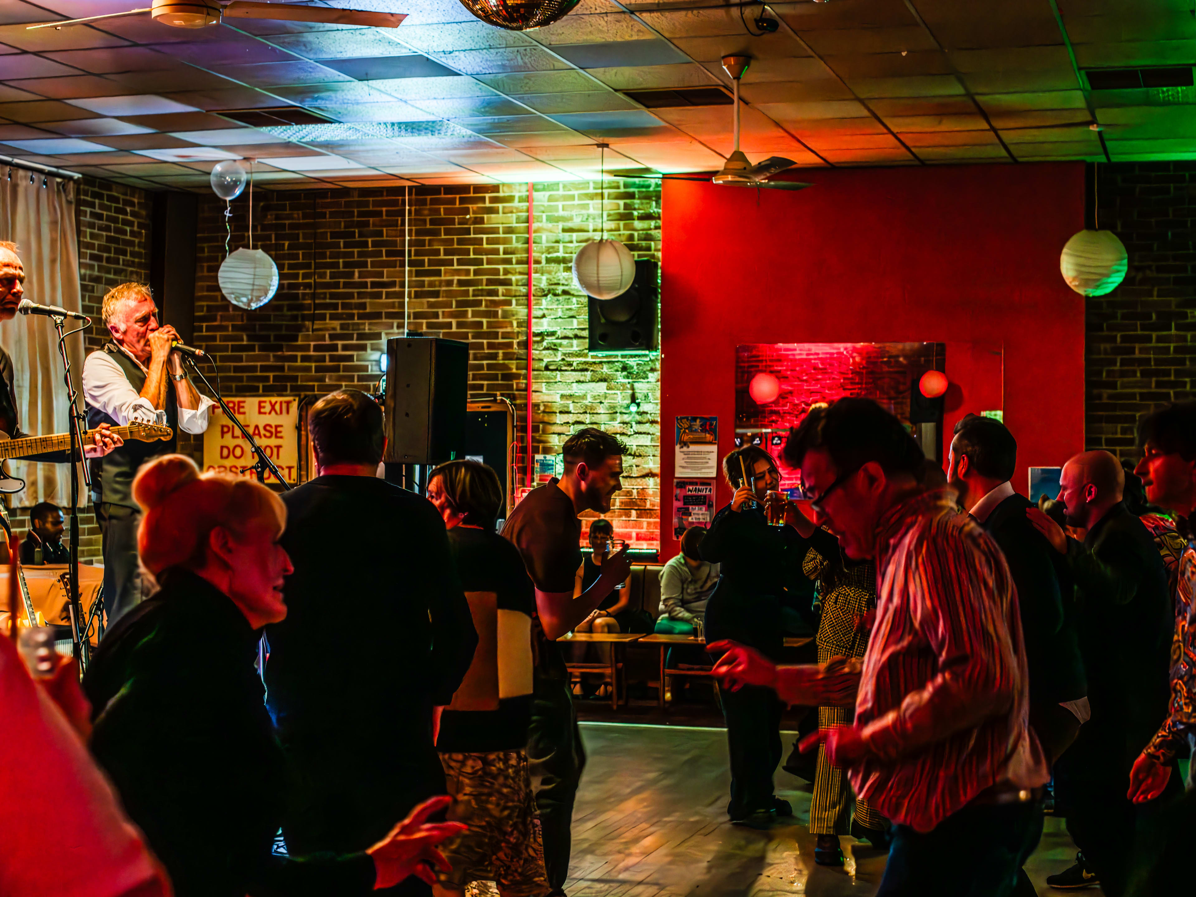 Live musicians singing and playing guitar on a stage. A crowd of people dance on the wooden dance floor at the red and green lit Walthamstow Trades Hall.