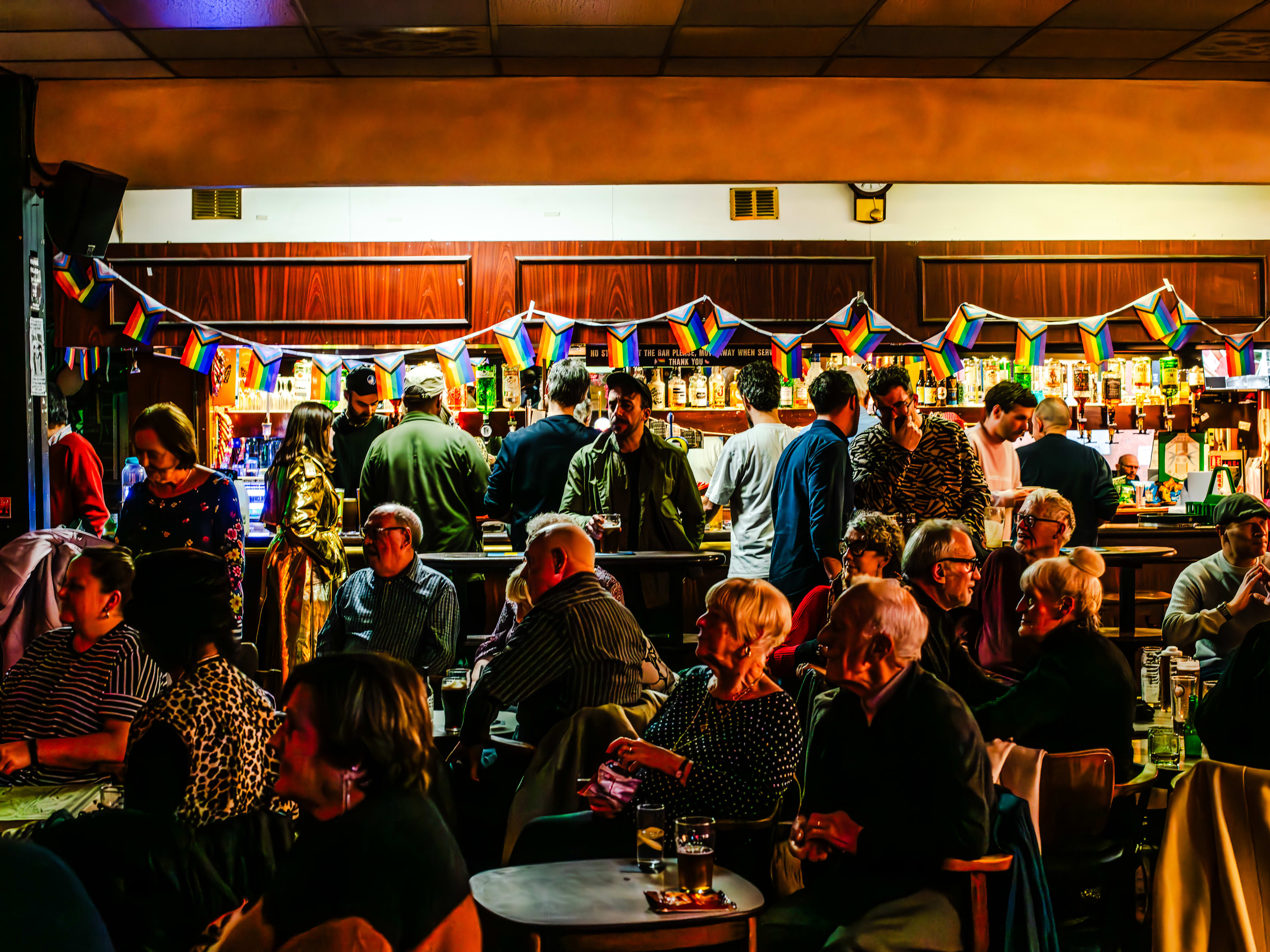 The crowded seating area at Walthamstow Trades Hall. Rainbow flags hang above the bar in the back where people are also standing and sitting.