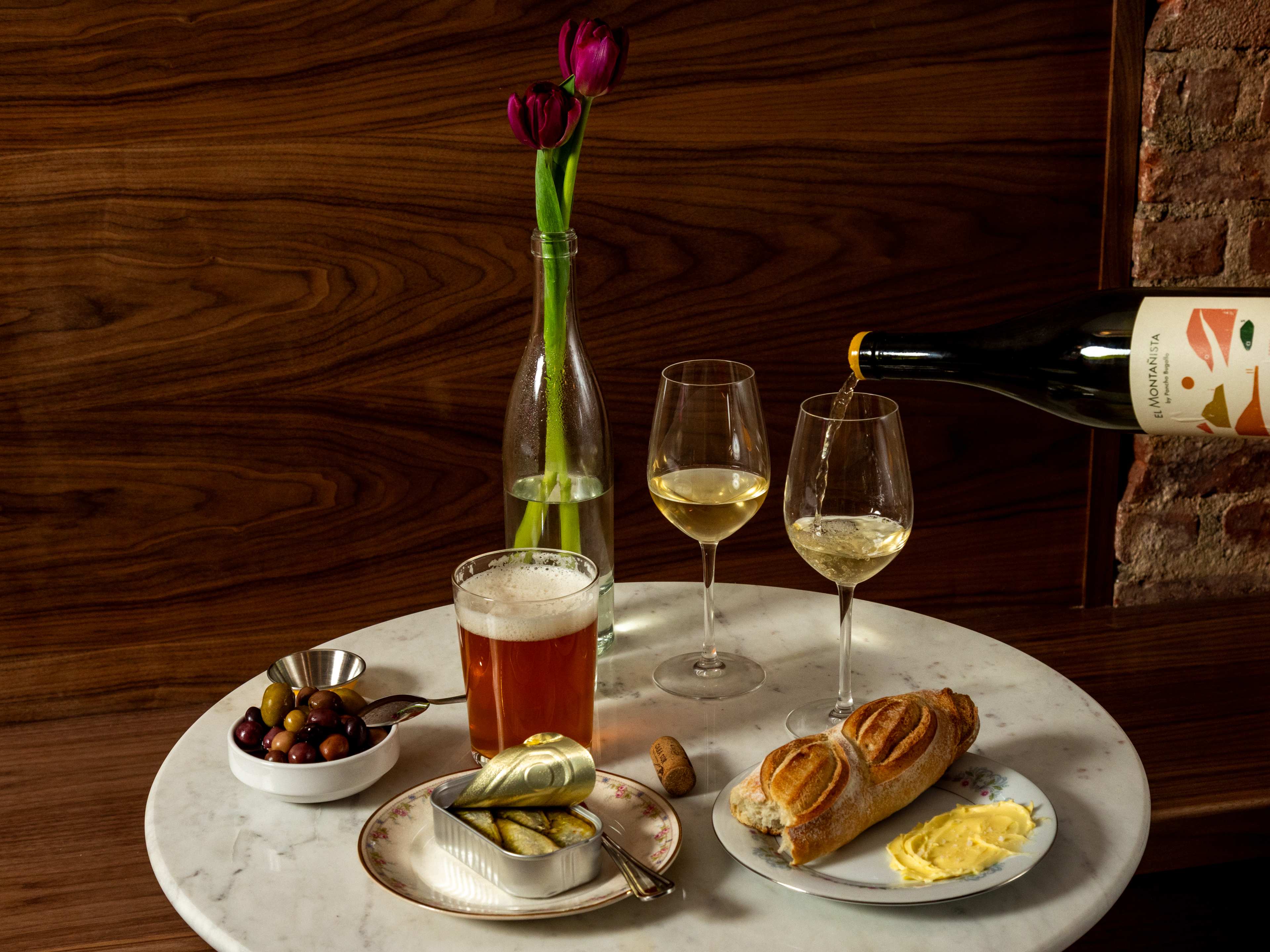 a table with olives, a beer, an opened can of sardines, a ripped baguette and butter, and two wine glasses