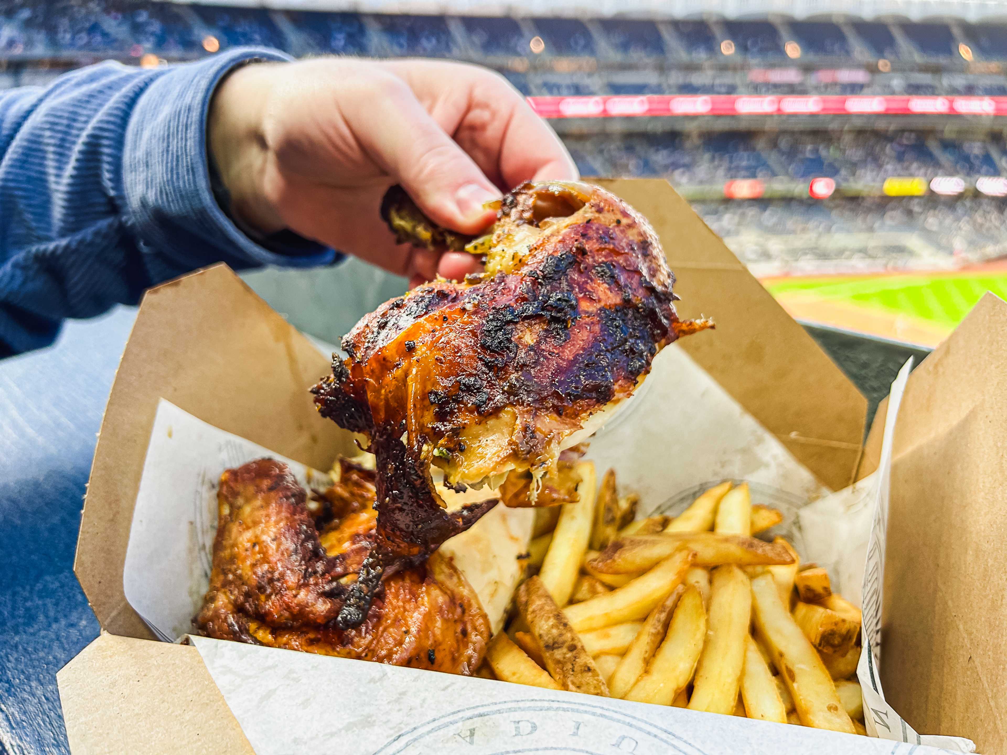 a hand holding up a chicken leg with a plate of fries with yankee stadium in the background