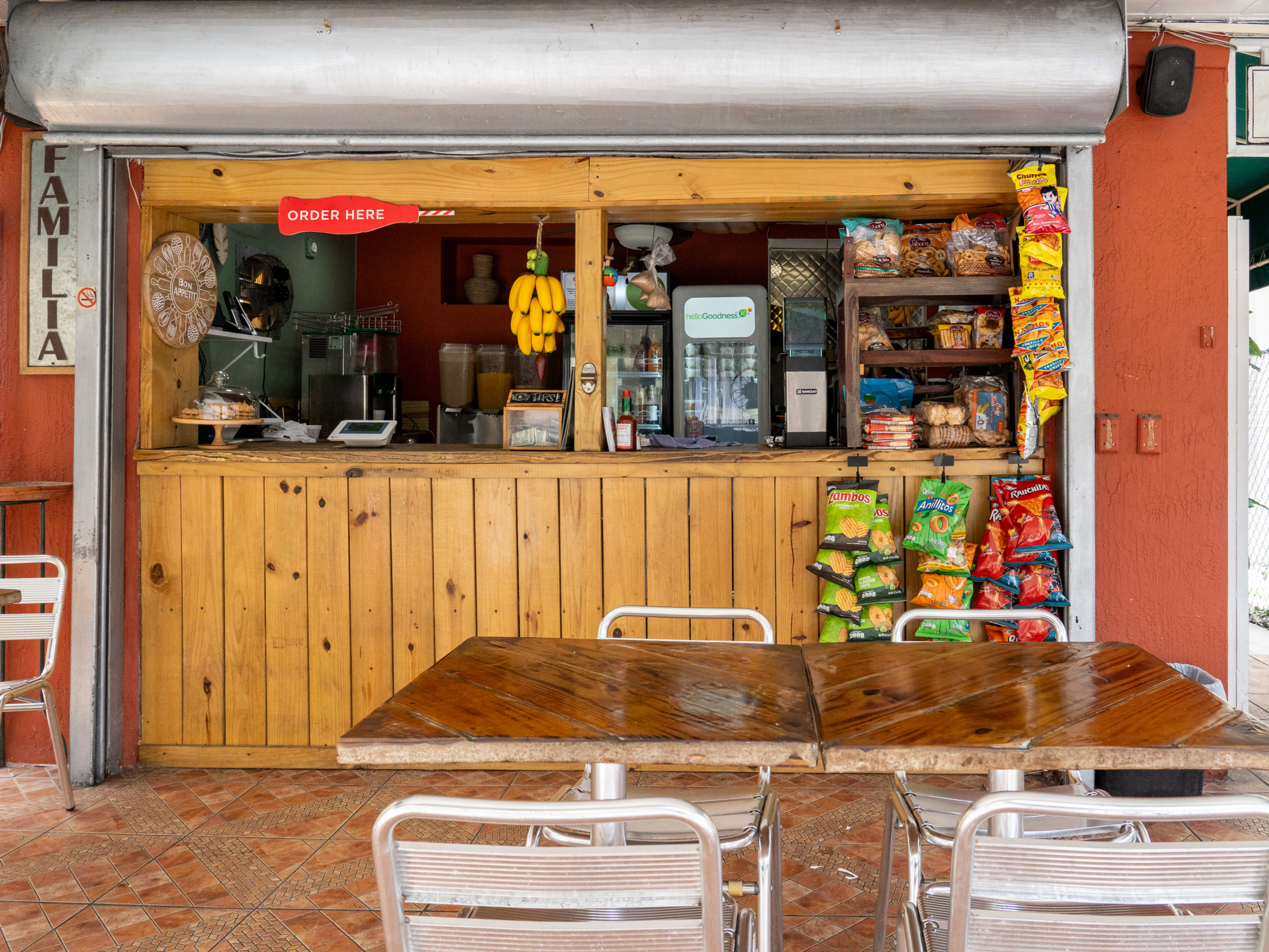 Outdoor restaurant with wood paneling, chips hanging on a stick, a vine of fake bananas, wooden tables, and metal chairs.