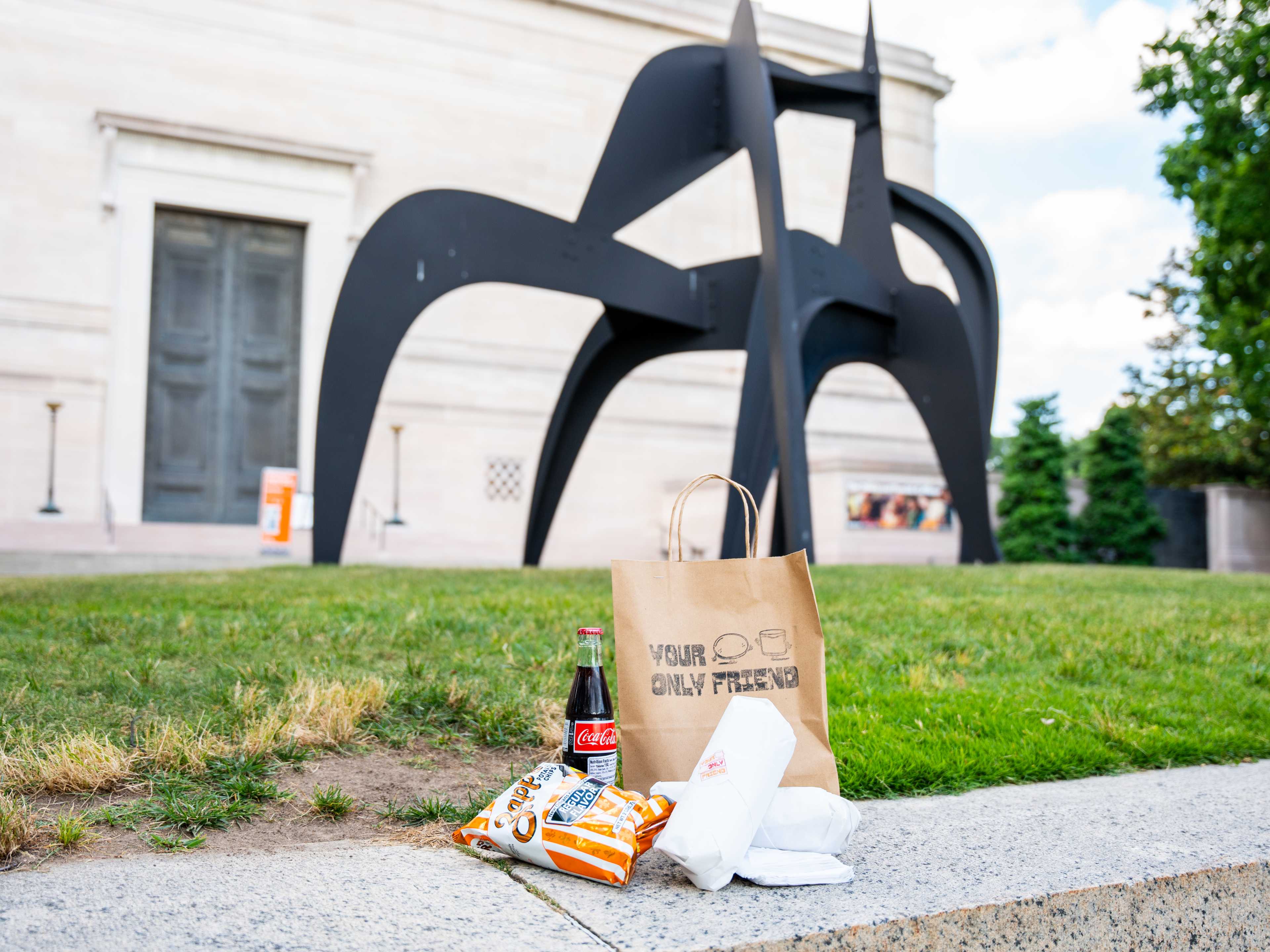 soda, sandwiches, and chips on ledge near massive outdoor abstract sculpture