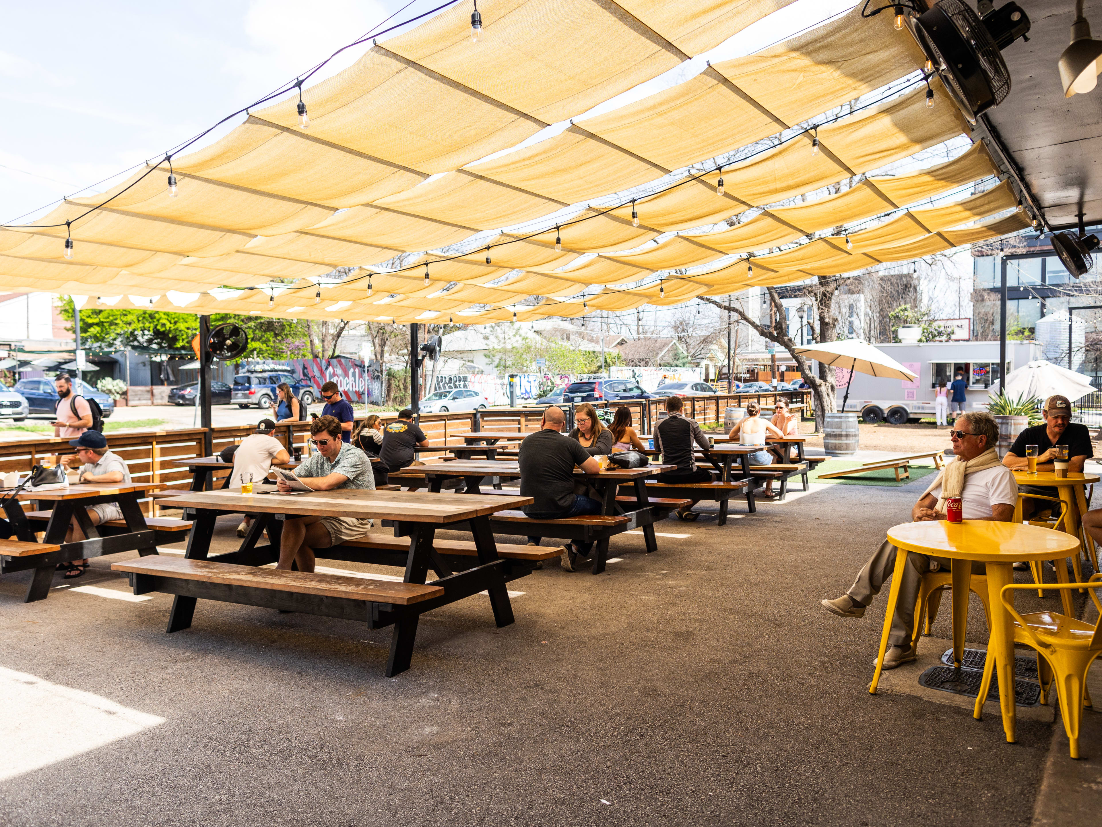 People sit at picnic tables, and yellow cafe tables under the canopy covered patio at Zilker Brewing.