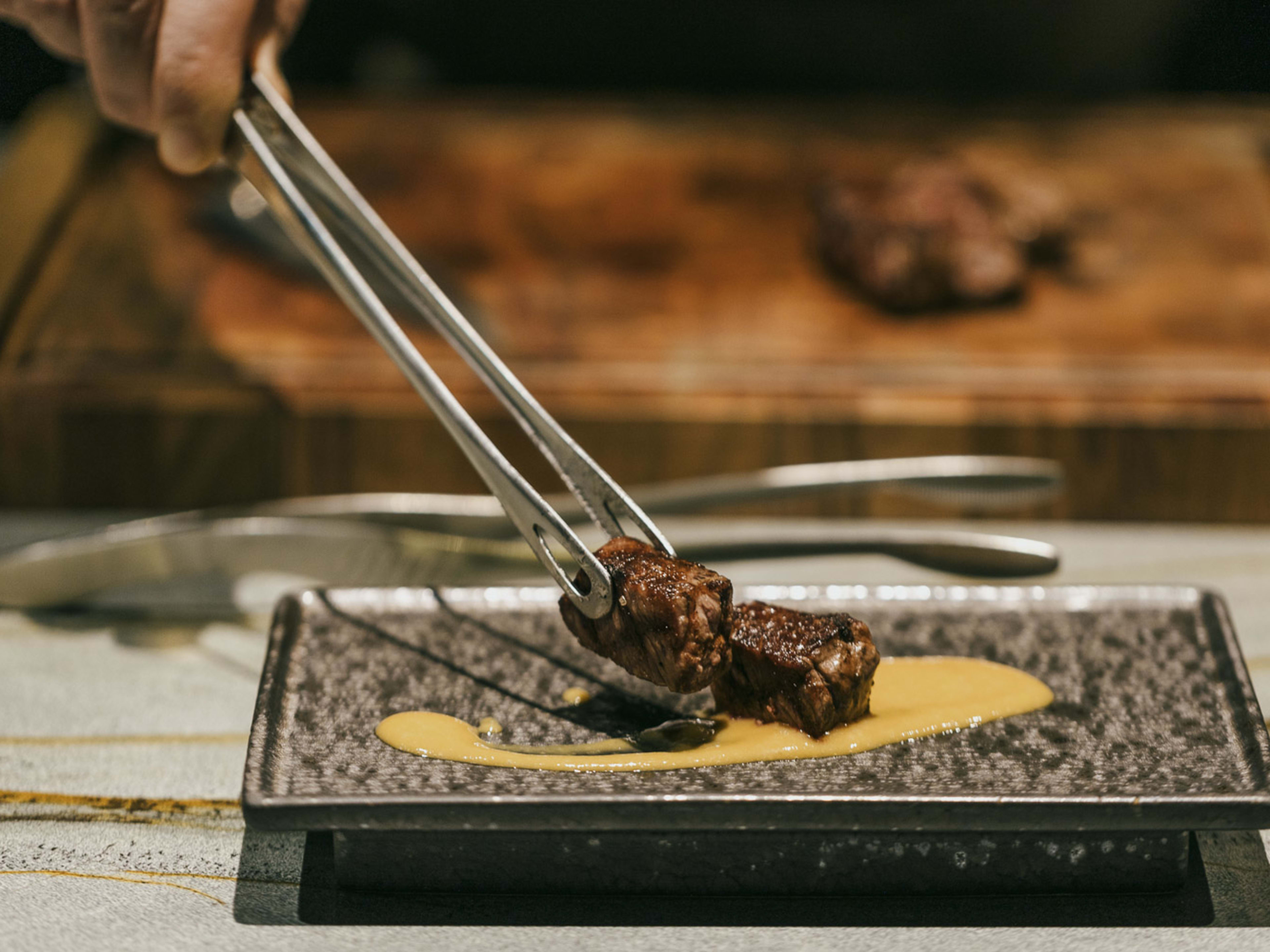 Tongs place two pieces of Ribeye with ssamjang puree on a ceramic dish.