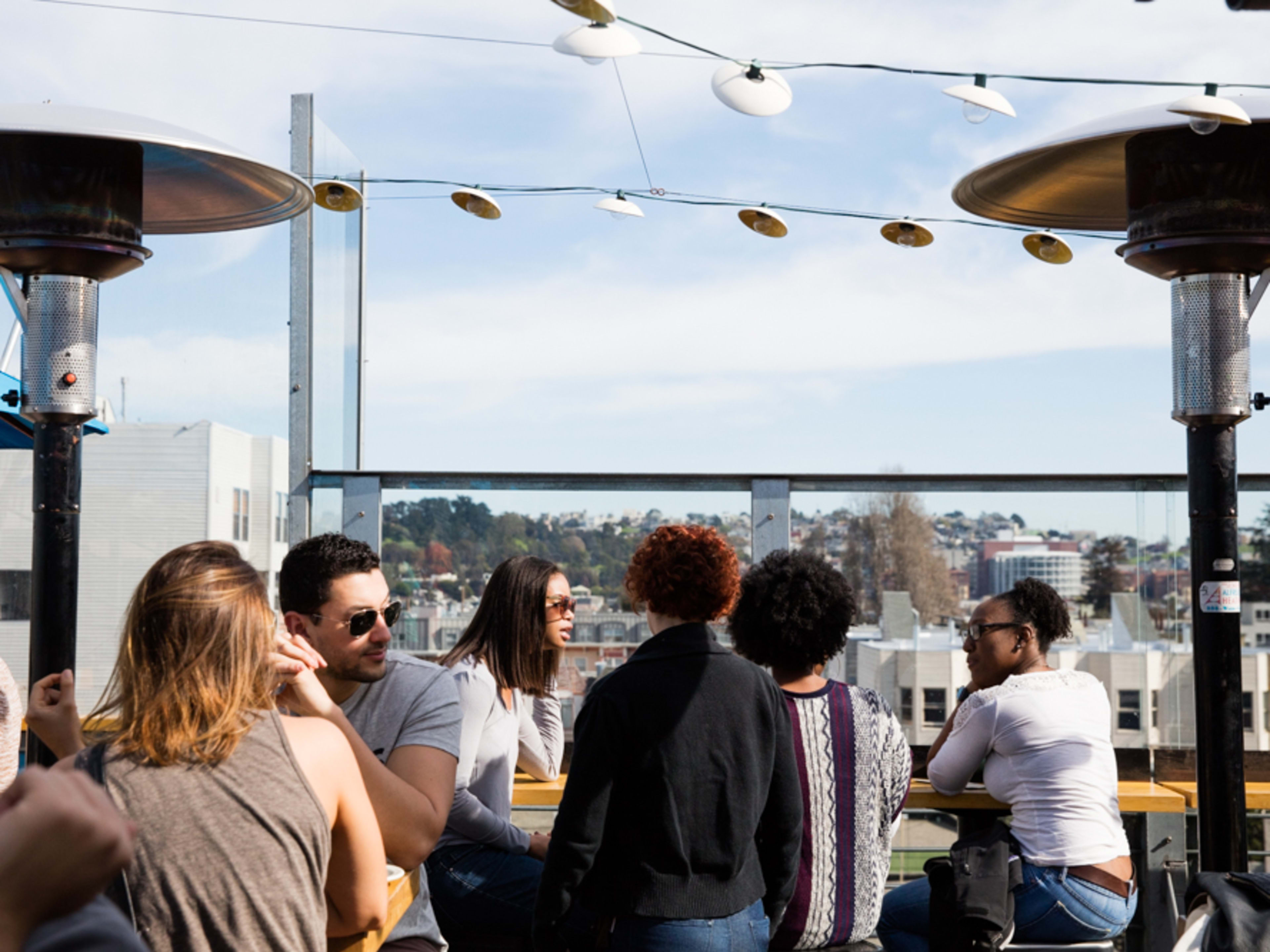 Sunny rooftop bar with heat lamps and string lights, filled with people.