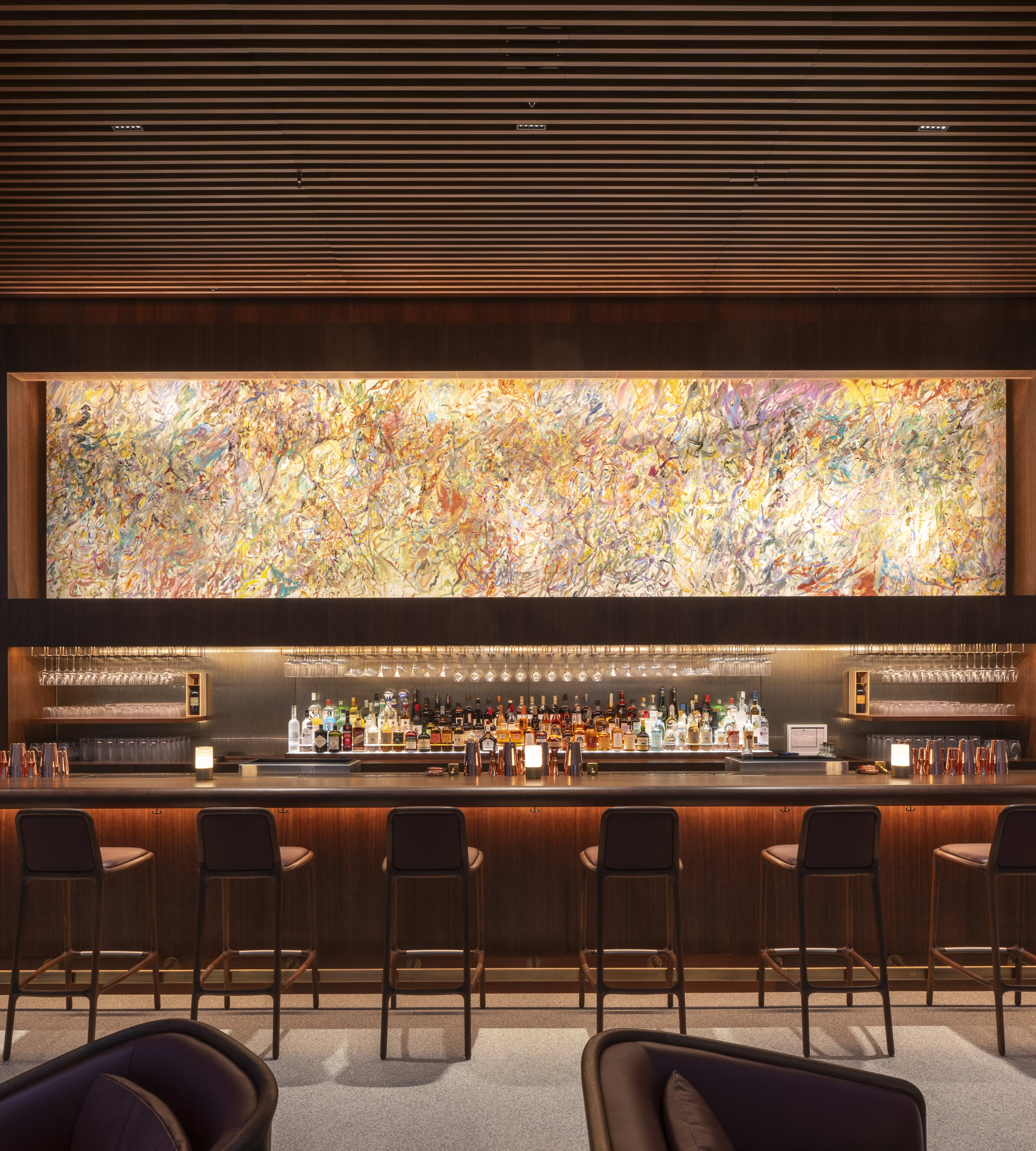 A long bar in the the bottom of a restaurant with a big illuminated abstract painting behind it.