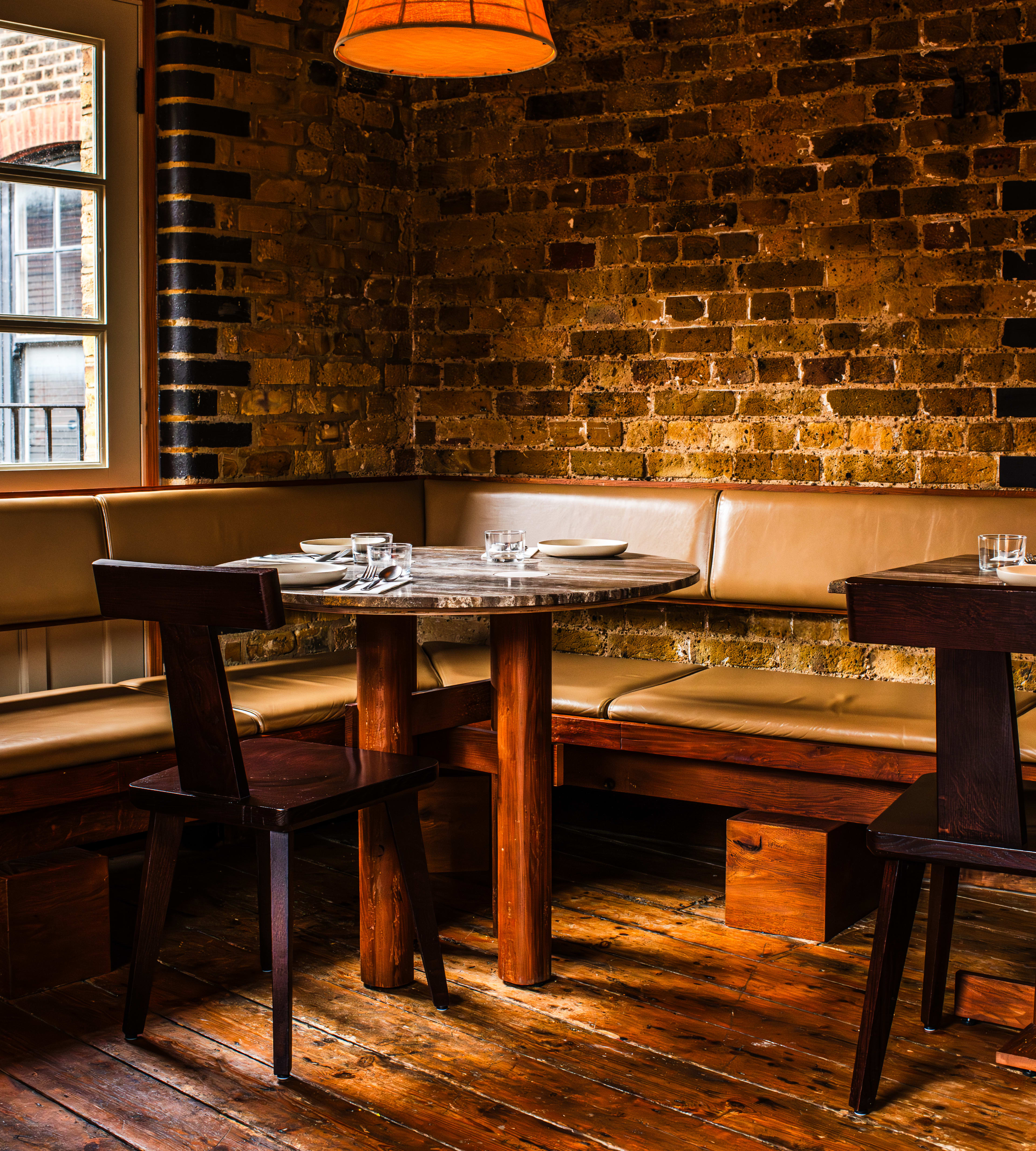 A long leather booth snakes around a corner at Kolae.  There are square tables with wooden chars, brick walls and a hanging pendant light.