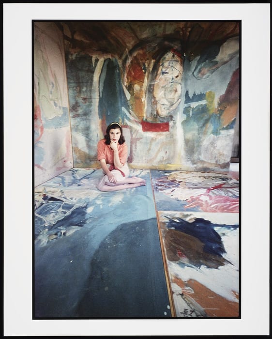 Photograph of the artist Helen Frankenthaler sitting on the floor surrounded by her own paintings.