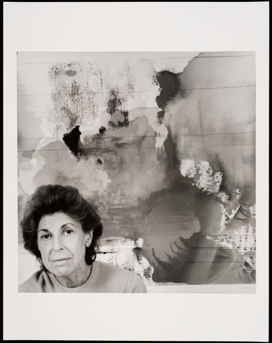 Black and white photograph. In the lower left corner a woman’s head and shoulders. She has dark hair and dark eyes. She is in front of a large abstract work of art with organic and overlapping shapes.