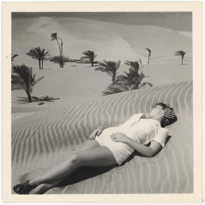 B&W photograph of a woman in a white romper lying on a hill of lined sand, hills and blowing palm trees in the background.