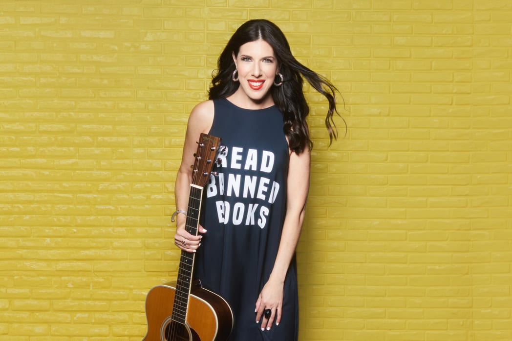 Joanie Leeds standing in front of a yellow wall with a shirt that says “read banned books” holding her guitar