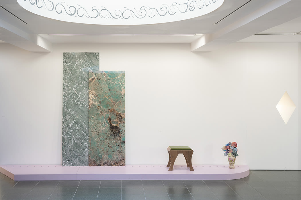 This image is an instillation shot of a white room with a lilac platform against the wall. From left to right on the platform are two large marble slabs leaning against the wall, a wooden stool with a green top, and a vase with pink and blue flowers.