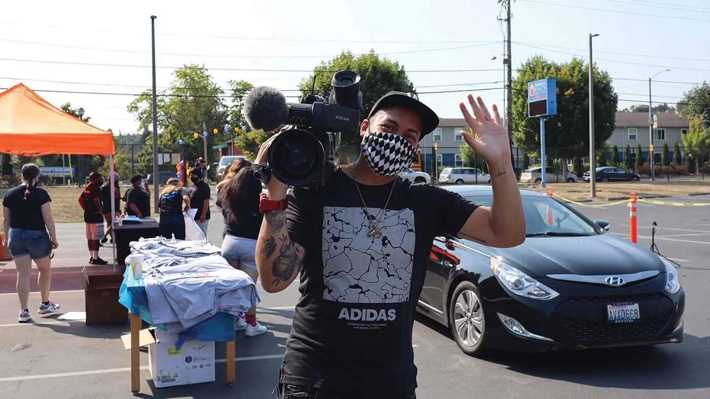 Man with black and white mask on waving, holding a filming camera with a car, people, and table behind him.