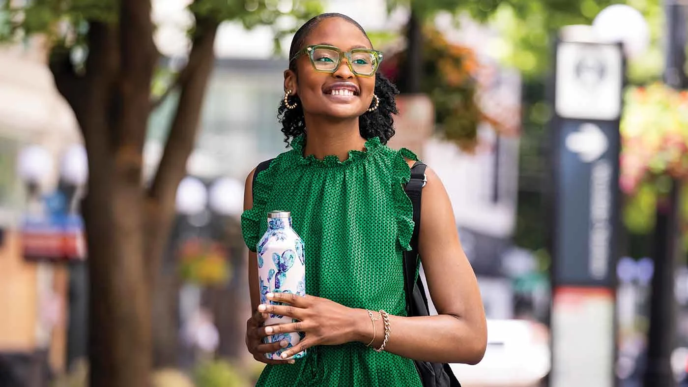 A young woman wearing a green dress, holding a water bottle walking down a city street.