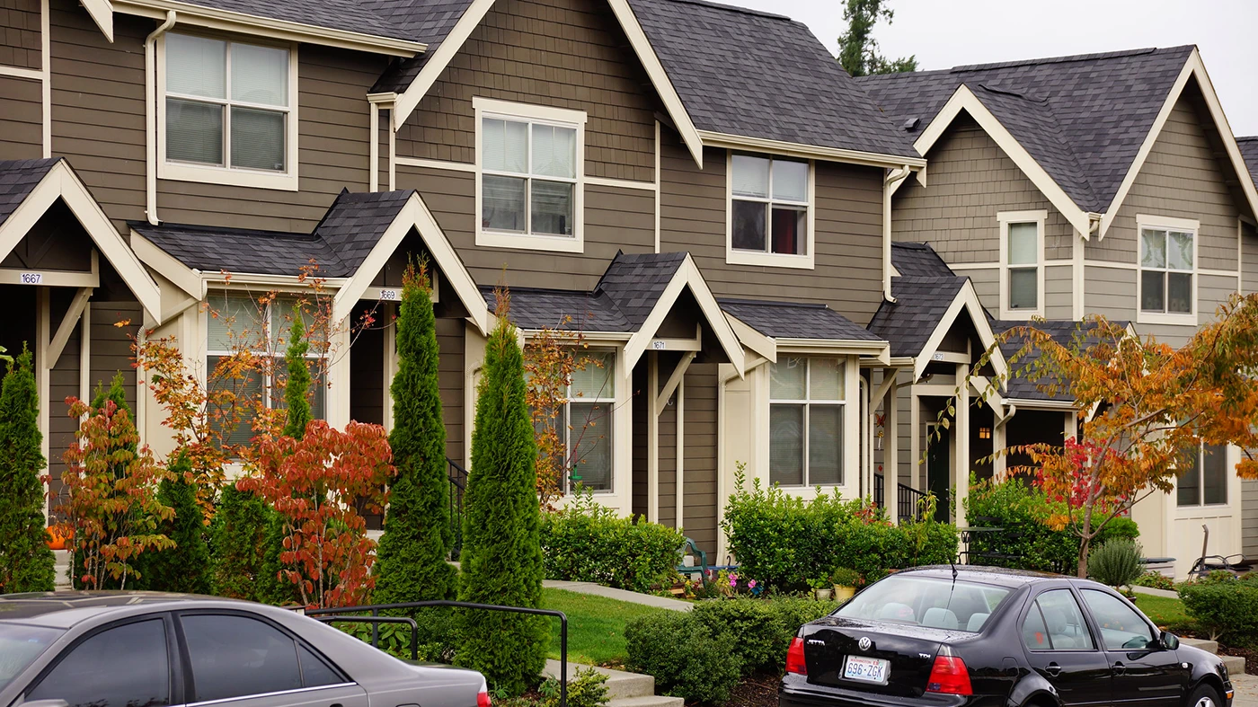 Brown houses with green lawn and trees and cars parked out front.